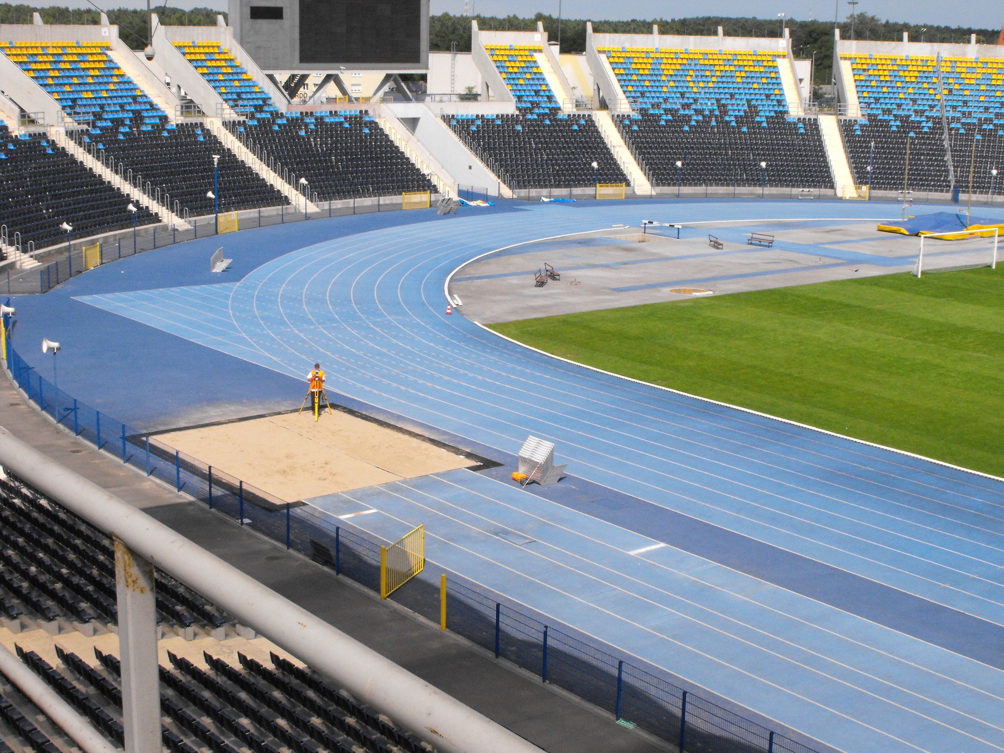 Pomiar geodezyjny na niebieskiej bieżni stadionu lekkoatletycznego, trybuny z żółto-czarnymi siedzeniami w tle.