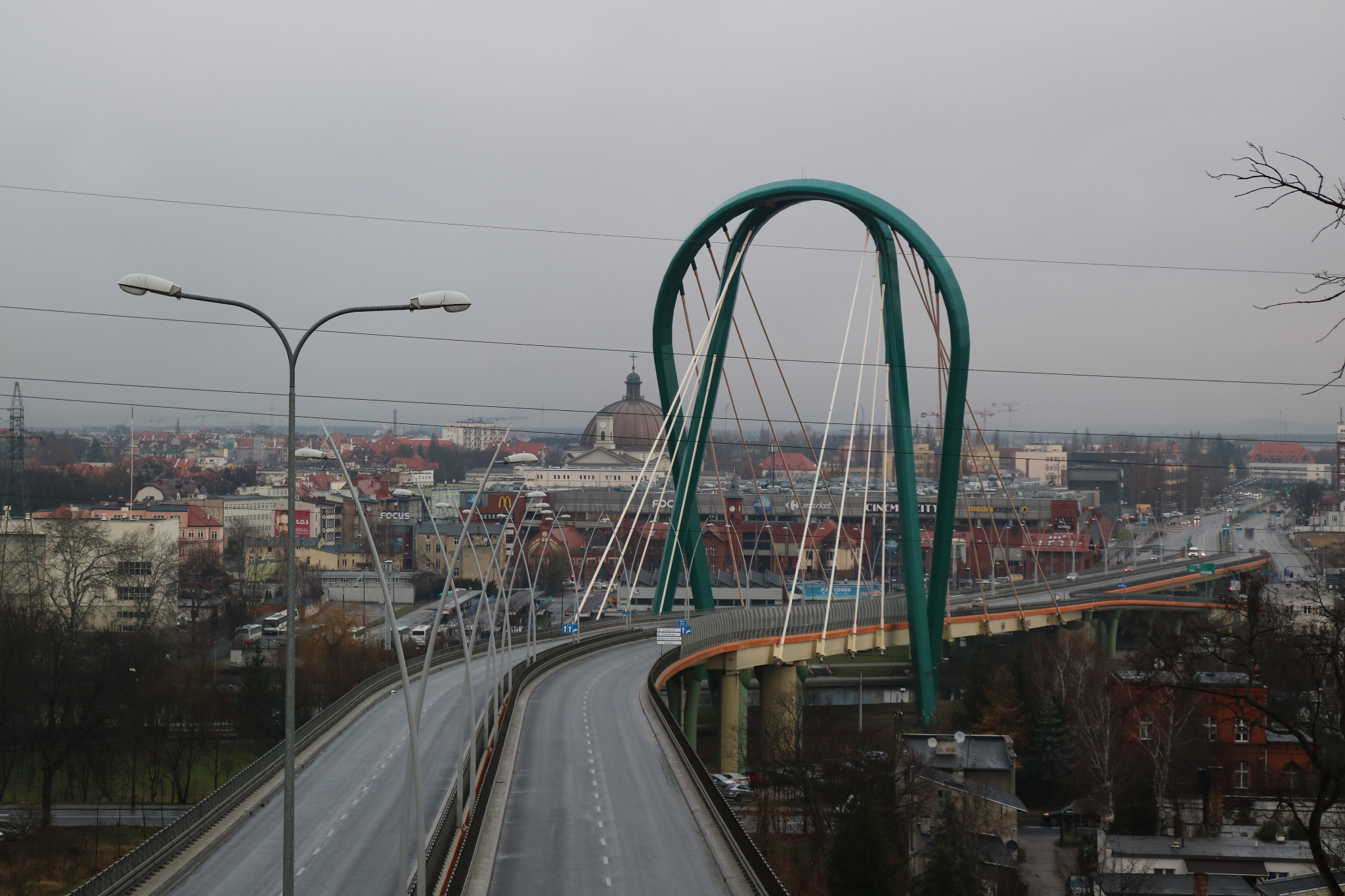 Most wantowy z zielonym łukiem w Grudziądzu, widok na miasto w pochmurny dzień, przewody elektryczne przecinają kadr.