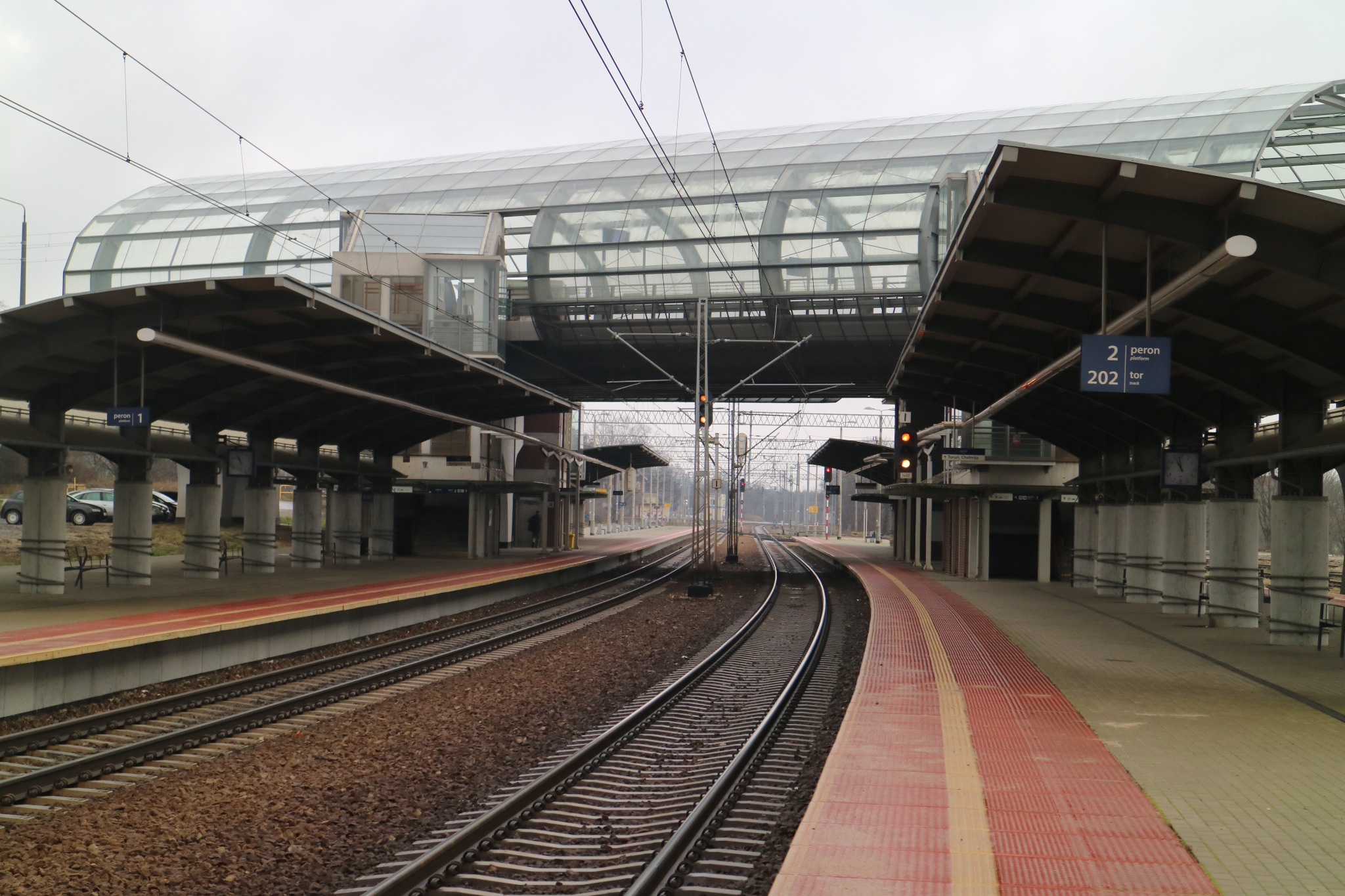Widok na perony kolejowe z zadaszeniem i szklanym łącznikiem dla pasażerów, widoczne tory i sygnalizacja świetlna.
