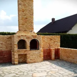 Murowany grill ogrodowy z cegły klinkierowej, z kominem i miejscem na drewno, na kamiennej posadzce, z widokiem na dom i żywopłot.