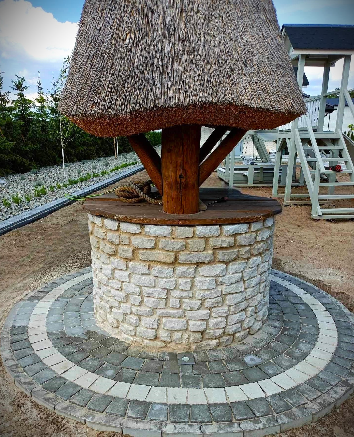 Studnia z kamienia wapiennego z drewnianym daszkiem krytym strzechą, otoczona koncentrycznymi kręgami z szaro-białej kostki brukowej, na tle placu zabaw i zieleni.