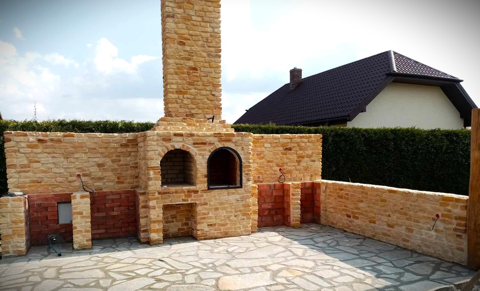 Murowany grill ogrodowy z cegły klinkierowej, z kominem i miejscem na drewno, na kamiennej posadzce, z widokiem na dom i żywopłot.