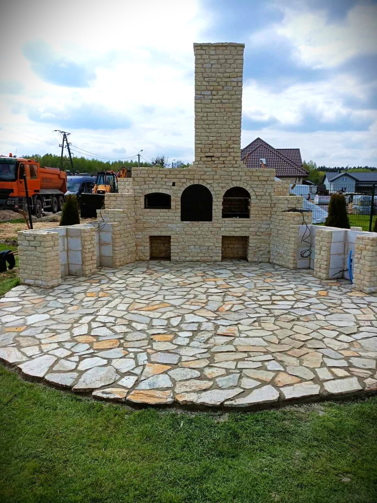 Kamienny taras z grillem i wędzarnią w trakcie budowy, z widocznymi elementami konstrukcyjnymi i materiałami budowlanymi, na tle zielonego trawnika i zabudowań mieszkalnych.