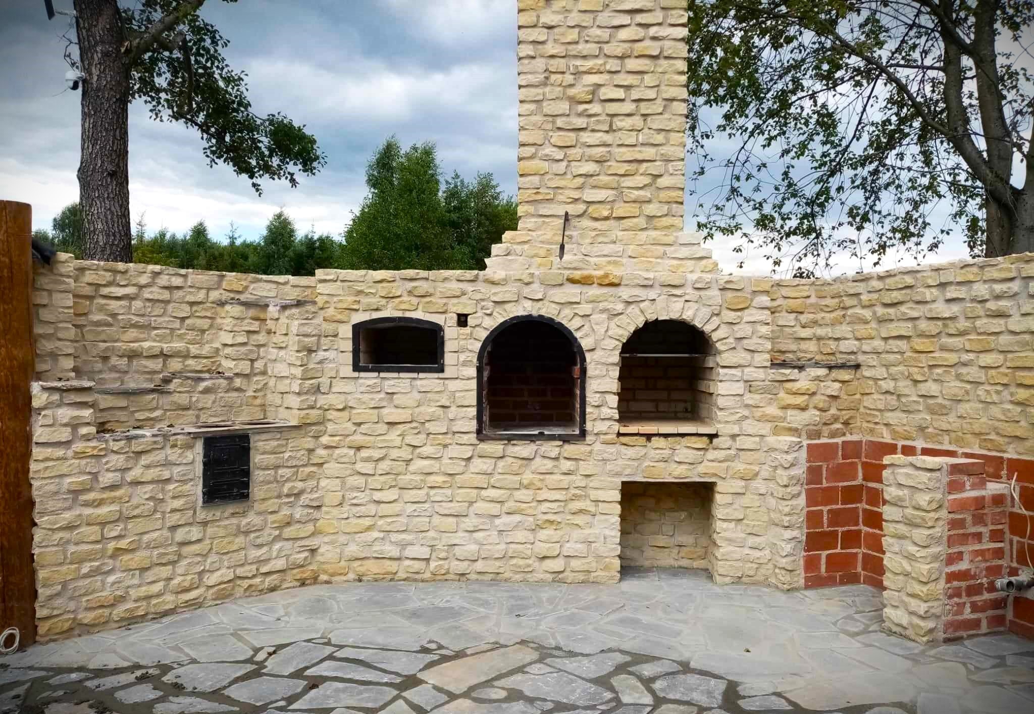 Murowana kuchnia letnia z kamienia i cegły, z otwartymi paleniskami i wędzarnią, na kamiennej posadzce, otoczona zielenią.