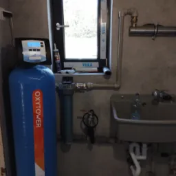 Kompletna instalacja uzdatniania wody w pomieszczeniu technicznym, widoczny niebieski zbiornik z napisem OxyTower, filtr narurowy, lampa UV, umywalka z plastikową butelką, okno z profilem VEKA.