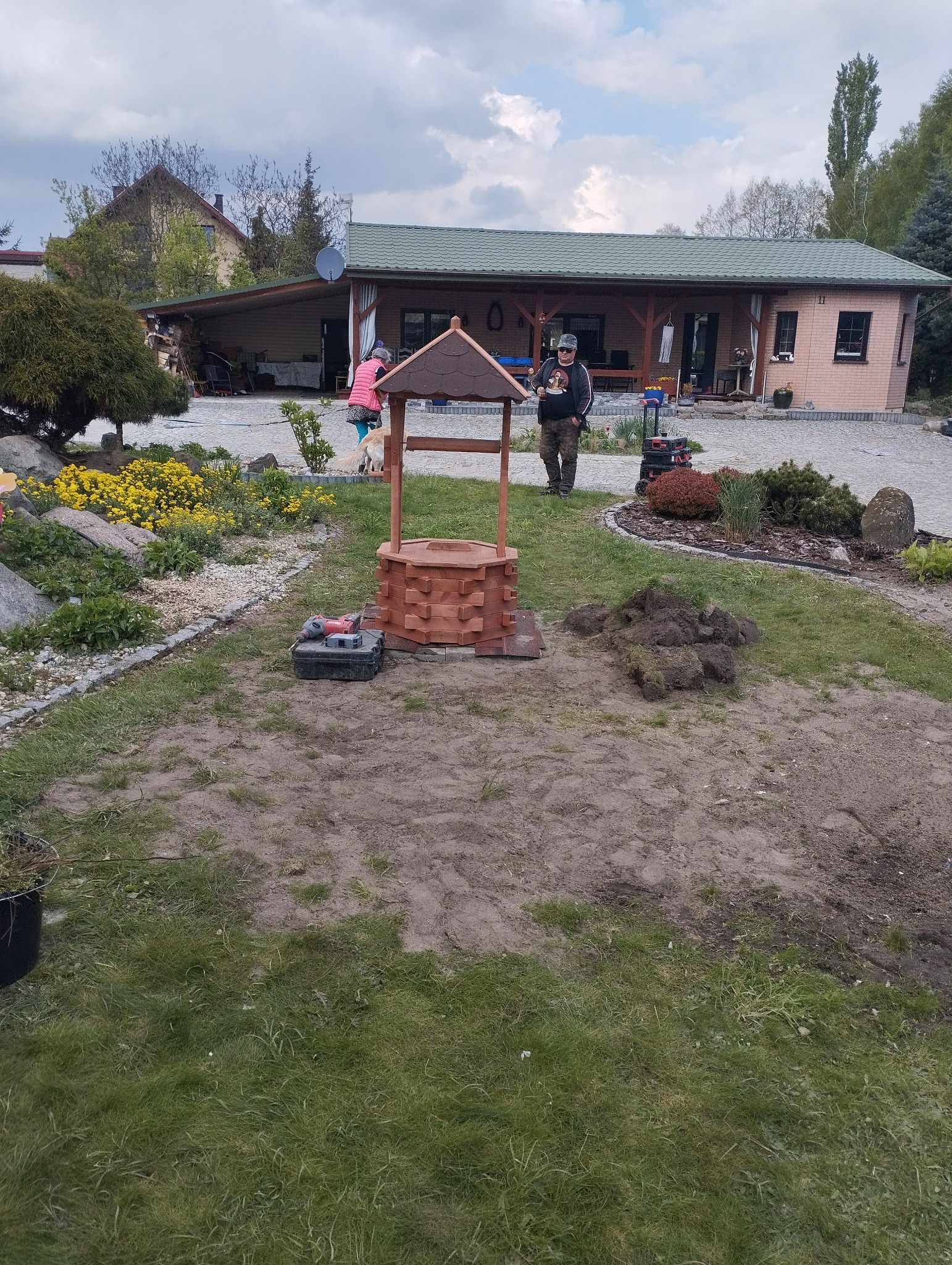 Drewniana studzienka dekoracyjna w ogrodzie, obok narzędzia, w tle dom z zielonym dachem i osoba z psem.