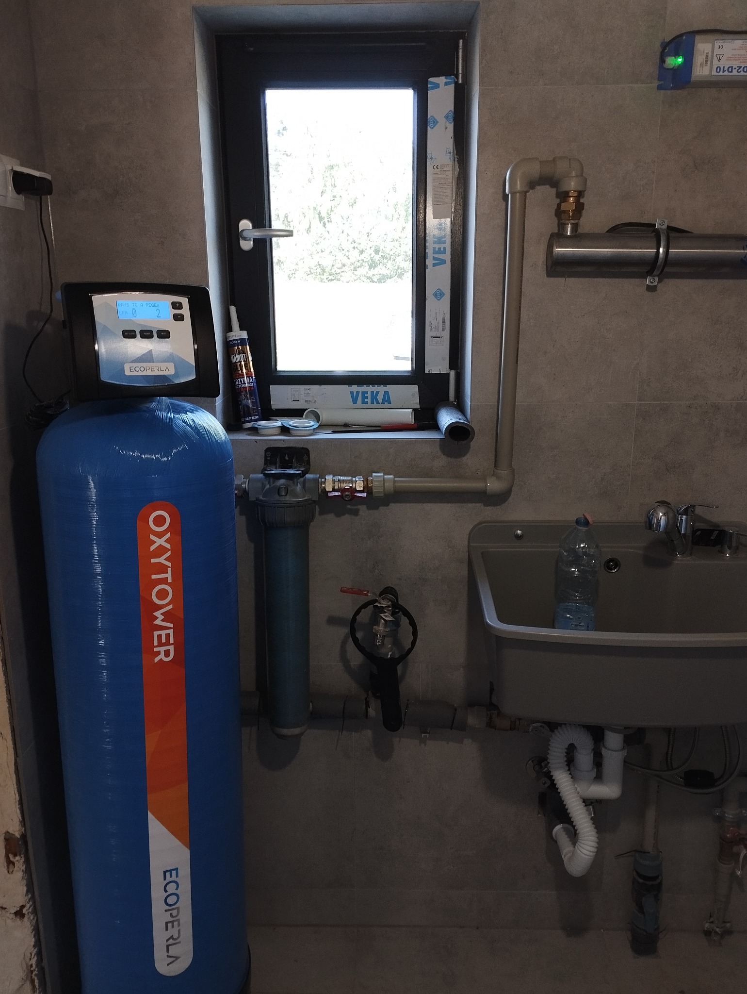 Kompletna instalacja uzdatniania wody w pomieszczeniu technicznym, widoczny niebieski zbiornik z napisem OxyTower, filtr narurowy, lampa UV, umywalka z plastikową butelką, okno z profilem VEKA.