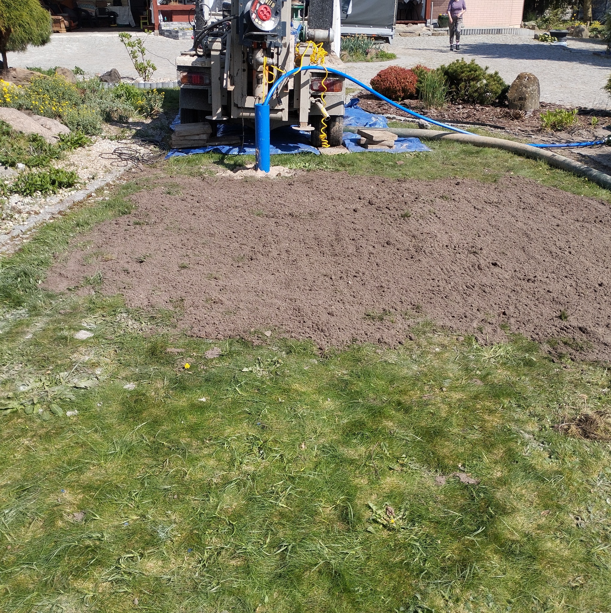 Wiertnica studni głębinowej na trawniku, widoczny wykop ziemi, niebieska rura wiertnicza i węże doprowadzające wodę.