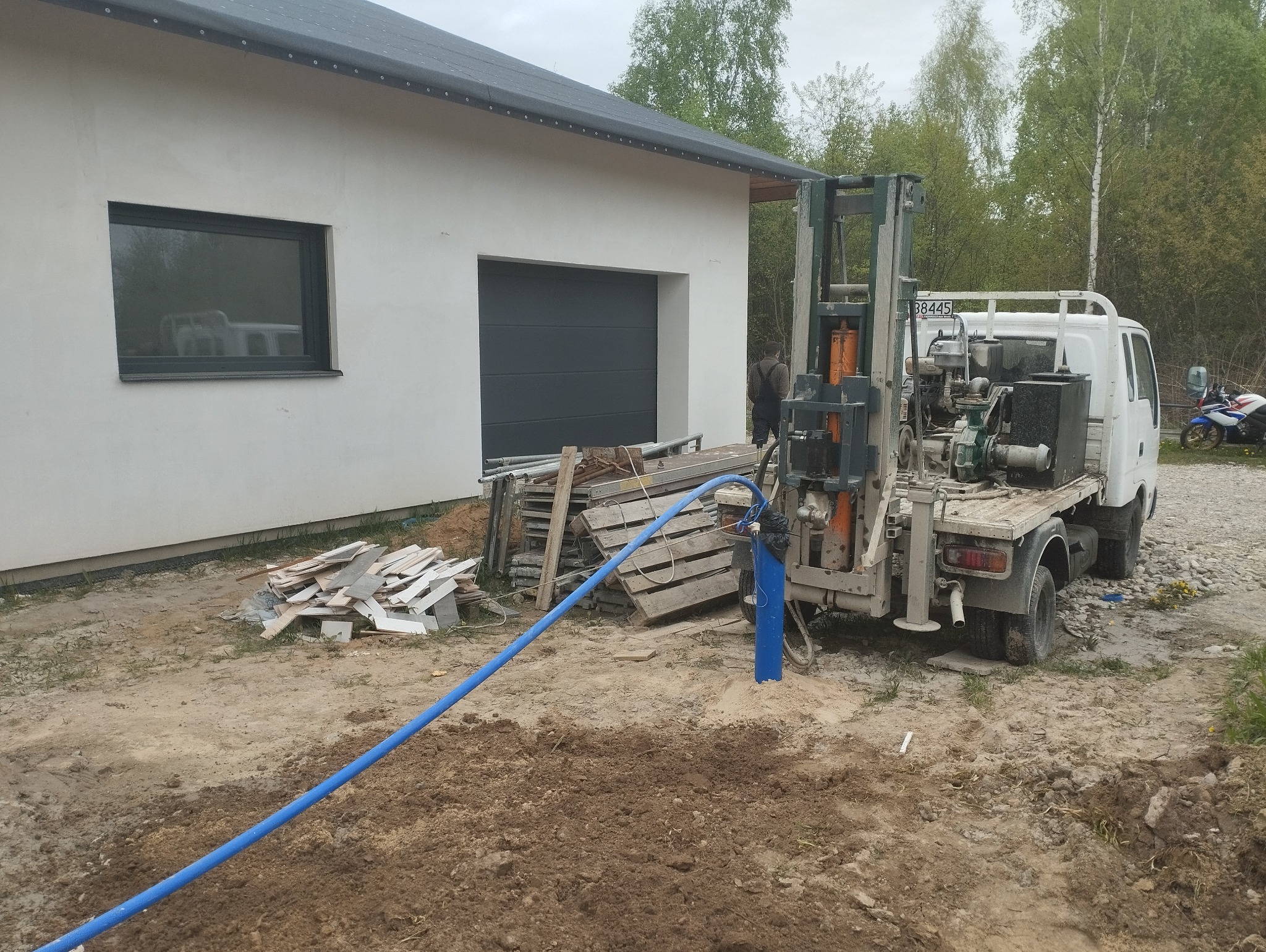Wiercenie otworu studziennego przy nowym domu, widoczna maszyna wiertnicza na samochodzie ciężarowym oraz niebieski wąż odprowadzający wodę.