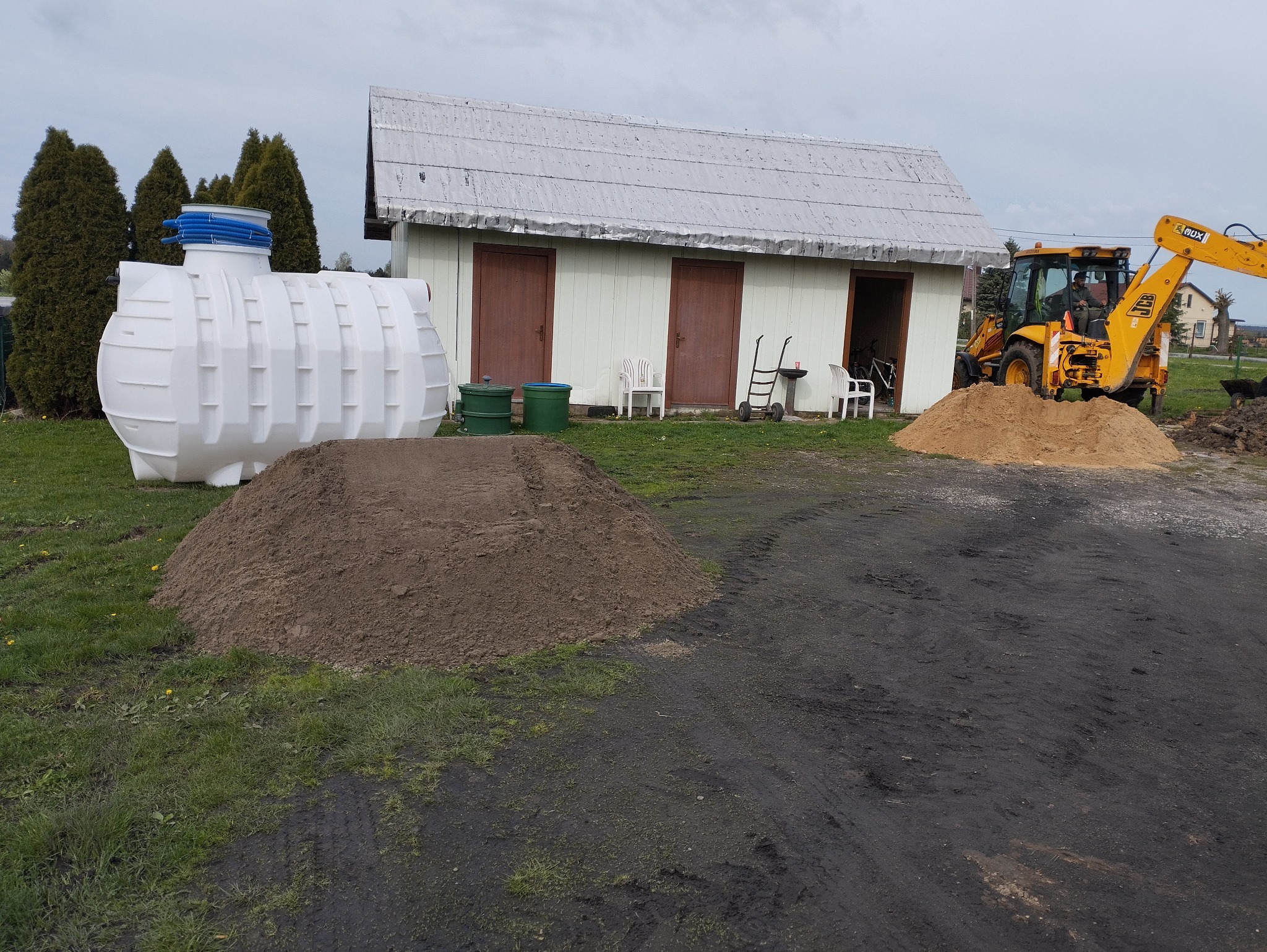 Biały zbiornik z niebieskimi rurami na trawie w Bełchatowie, obok pryzma ziemi i koparka JCB podczas prac ziemnych, w tle budynek gospodarczy i drzewa.