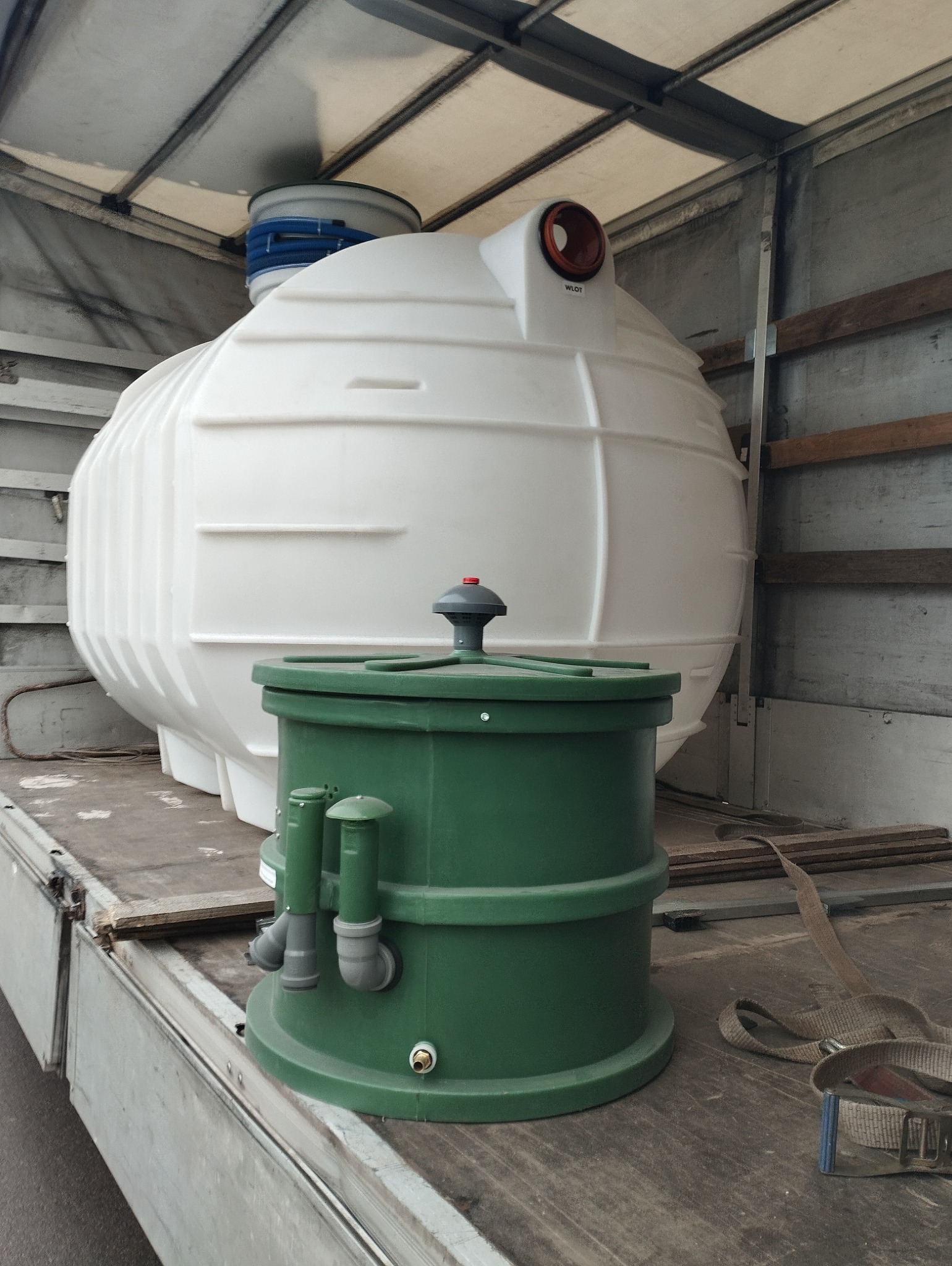 Biały zbiornik retencyjny i zielony separator tłuszczu w przestrzeni ładunkowej samochodu dostawczego, zabezpieczone pasami transportowymi.