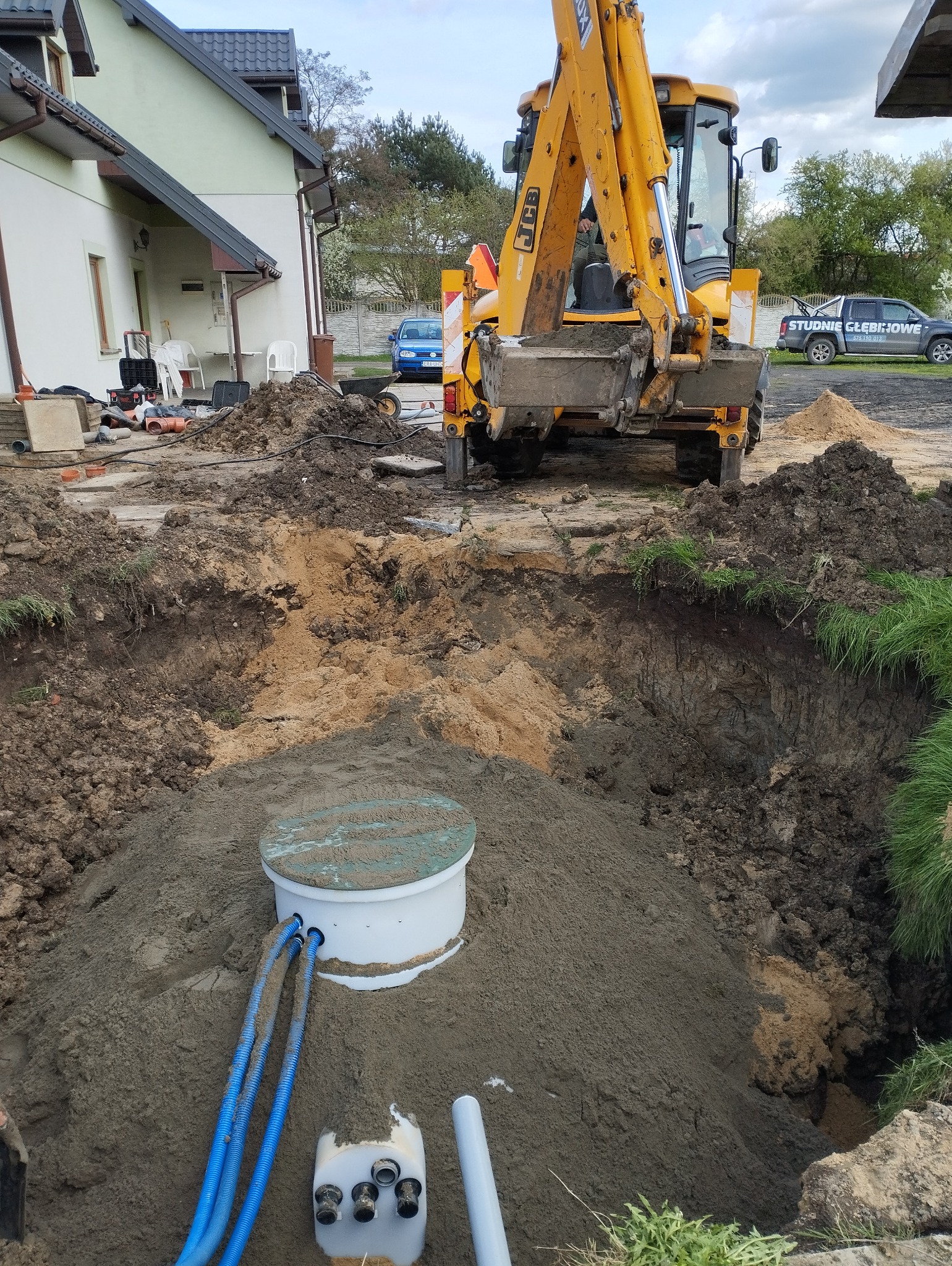 Żółta koparko-ładowarka zasypuje wykop z zamontowanym białym zbiornikiem z niebieskimi rurami w pobliżu domu jednorodzinnego.