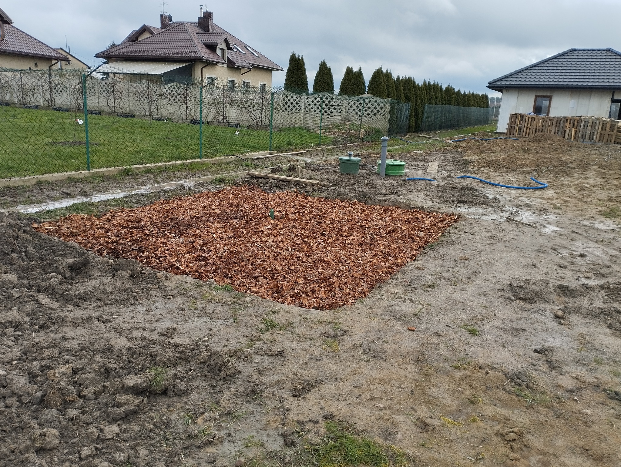 Kwadratowy wykop wypełniony brązowymi zrębkami drewna, widoczne dwa zielone włazy do zbiorników, w tle domy jednorodzinne i tuje, pochmurny dzień.