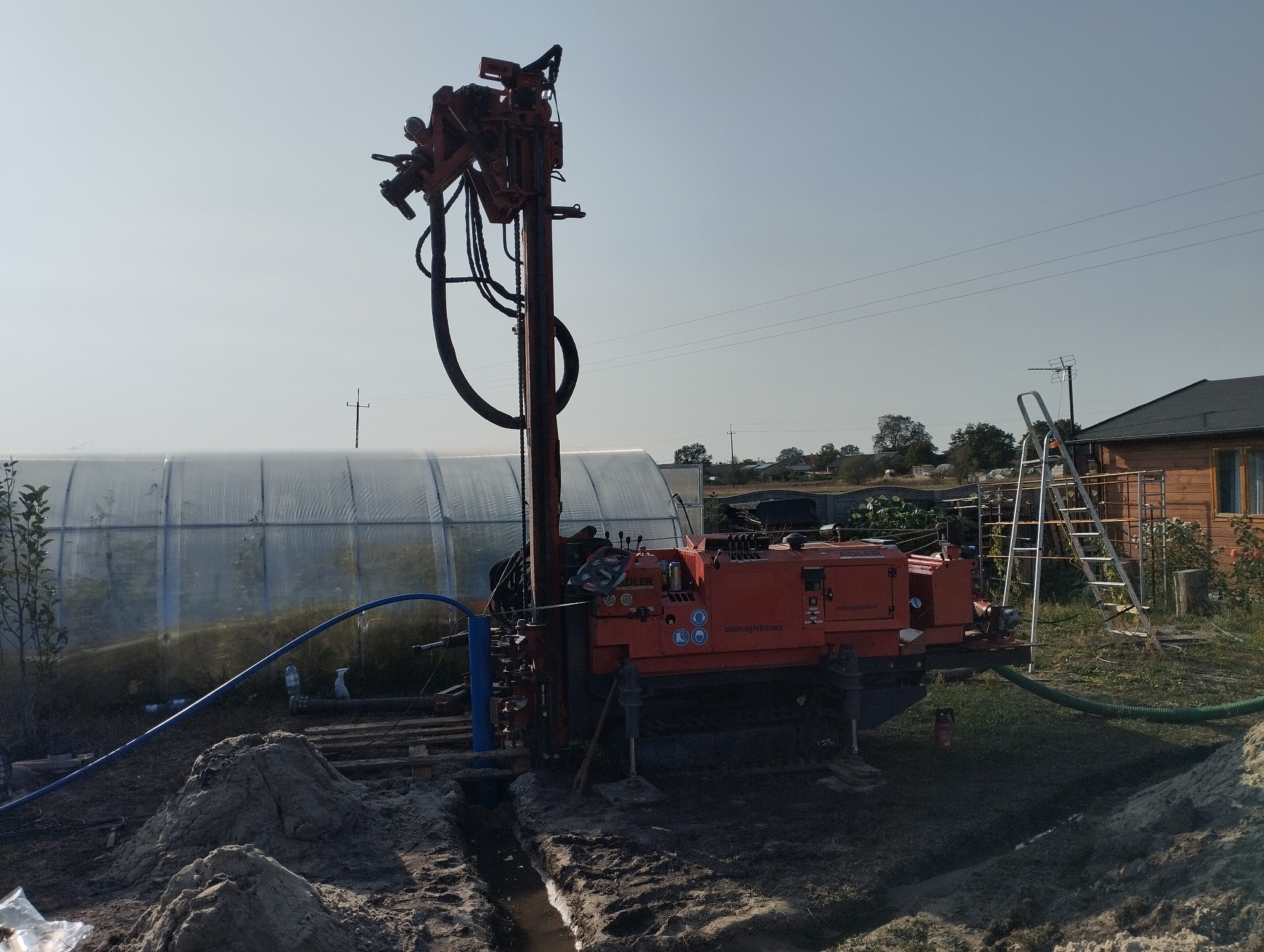 Pomarańczowa wiertnica na podwórku, widoczny fragment szklarni i drabina oparta o drewniany budynek w tle.