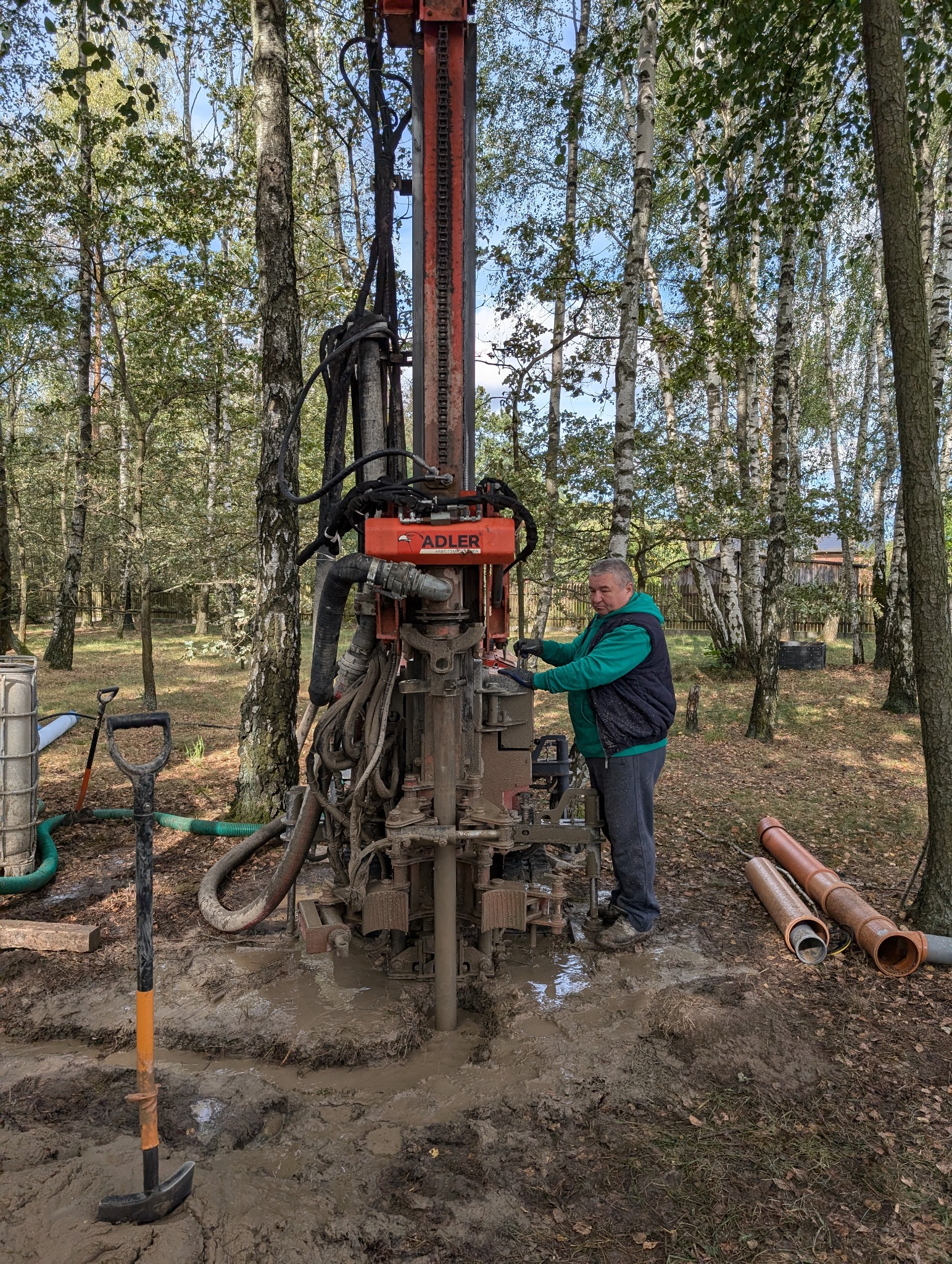Operator w zielonej bluzie obsługuje wiertnicę Adler w błotnistym terenie leśnym, widoczne rury i szpadel.