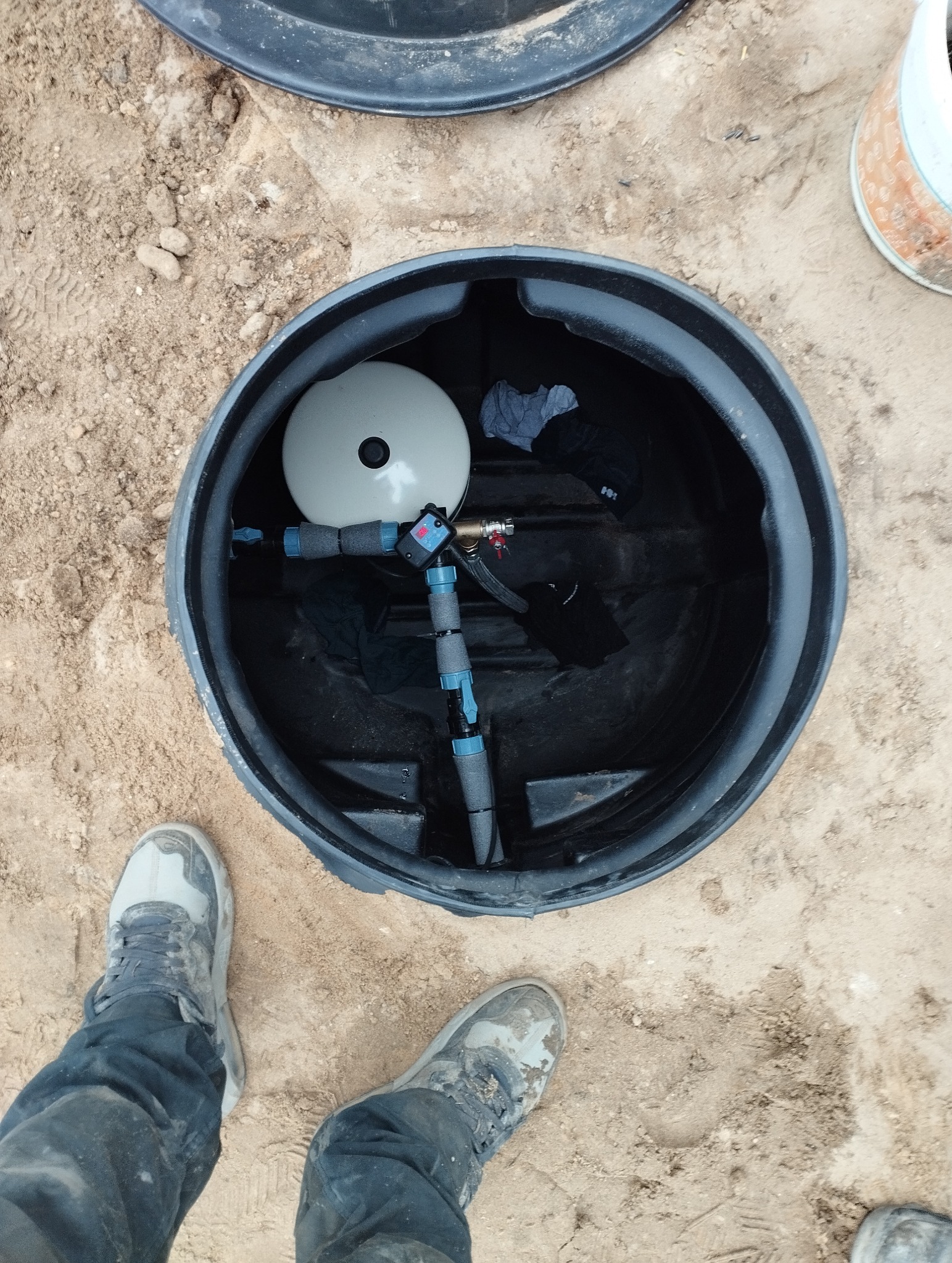 Wnętrze czarnego zbiornika z szarą membraną hydroforową, rurami z niebieskimi zaworami i czarnymi akcesoriami, widoczne z góry, z fragmentem butów na piaszczystym terenie.