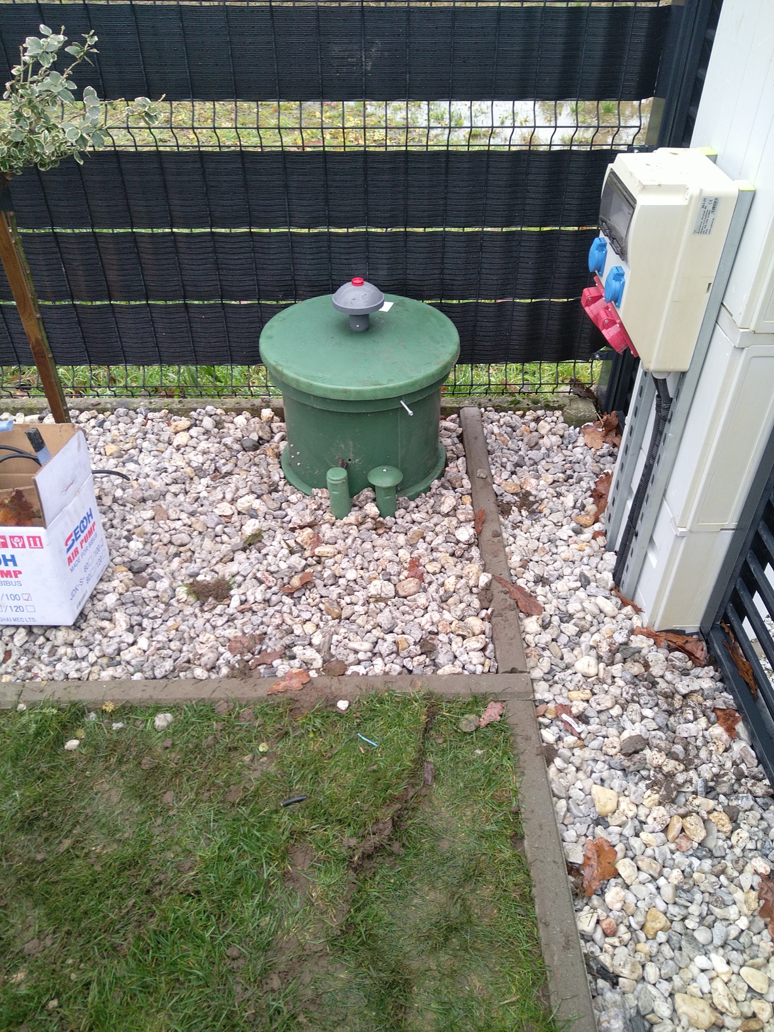 Zielony zbiornik z szarą pokrywą i czerwonym elementem, prawdopodobnie część instalacji oczyszczania ścieków, otoczony kamieniami i fragmentem trawnika, obok skrzynka elektryczna z gniazdami.