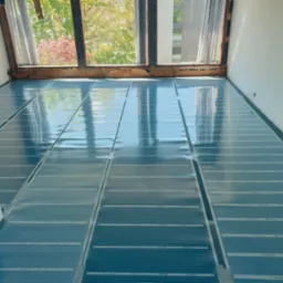 Rozłożone, przezroczyste panele systemu ogrzewania podłogowego na podłożu, widok z góry, okno z widokiem na drzewa w tle.