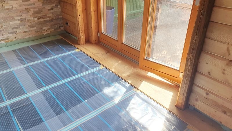 Montaż maty grzewczej na podłodze w drewnianym domu, widoczne niebieskie linie grzewcze i folia ochronna przed wylewką, drewniane ramy okienne w tle.