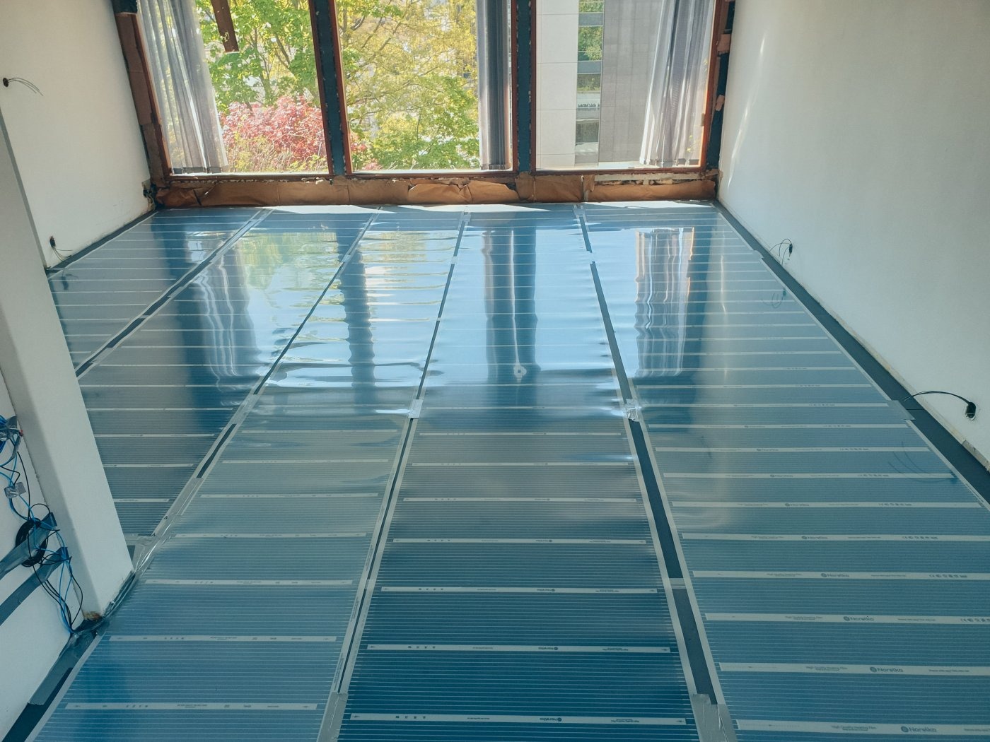 Rozłożone, przezroczyste panele systemu ogrzewania podłogowego na podłożu, widok z góry, okno z widokiem na drzewa w tle.