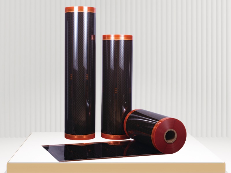 Zestaw trzech rolek folii grzewczej z miedzianymi akcentami, jedna rozwinięta na białym blacie, ukazująca strukturę węglową.