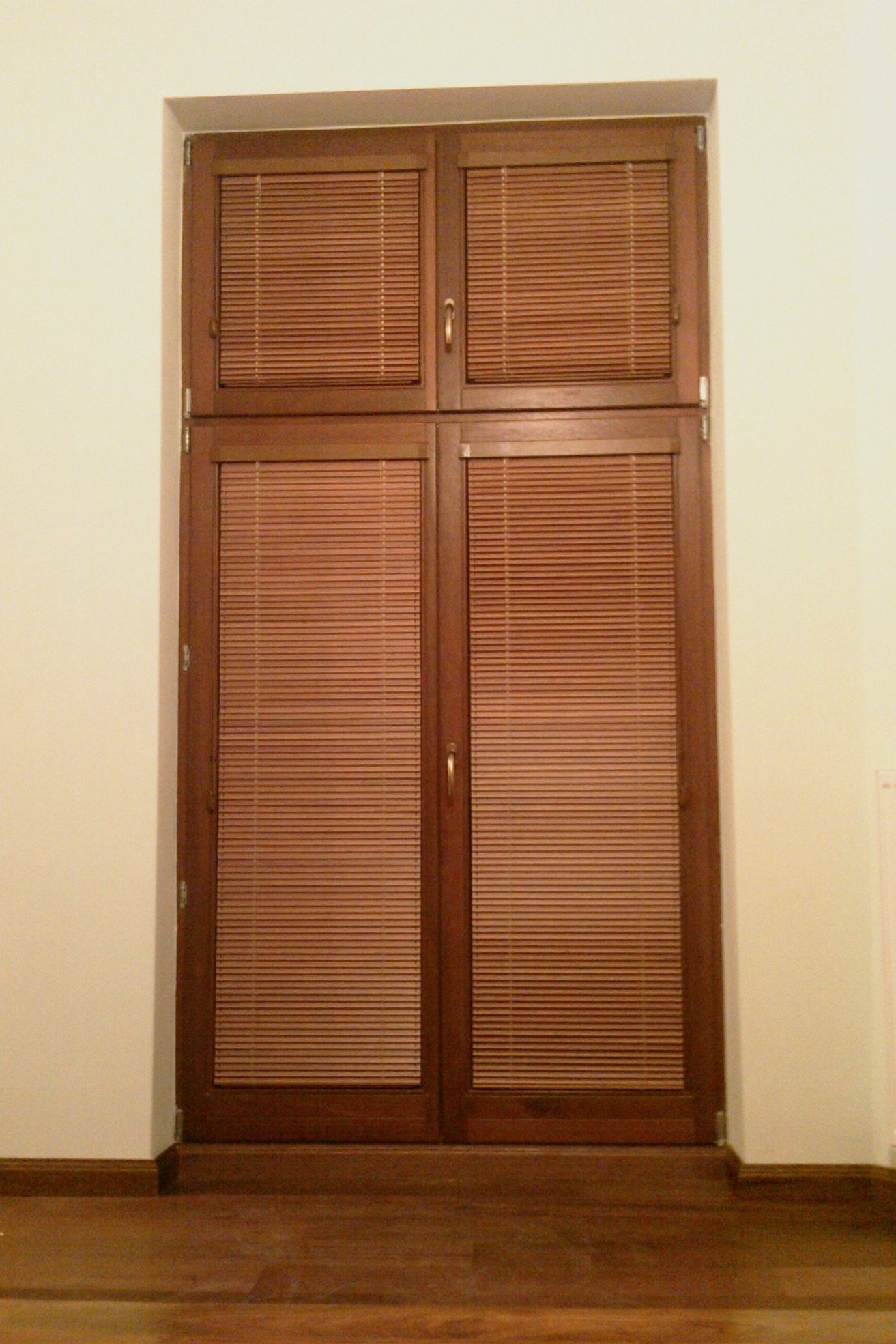 Drewniane okno z zamontowanymi, zamkniętymi żaluzjami w ciepłym odcieniu brązu, widoczne na tle jasnej ściany i drewnianej podłogi.