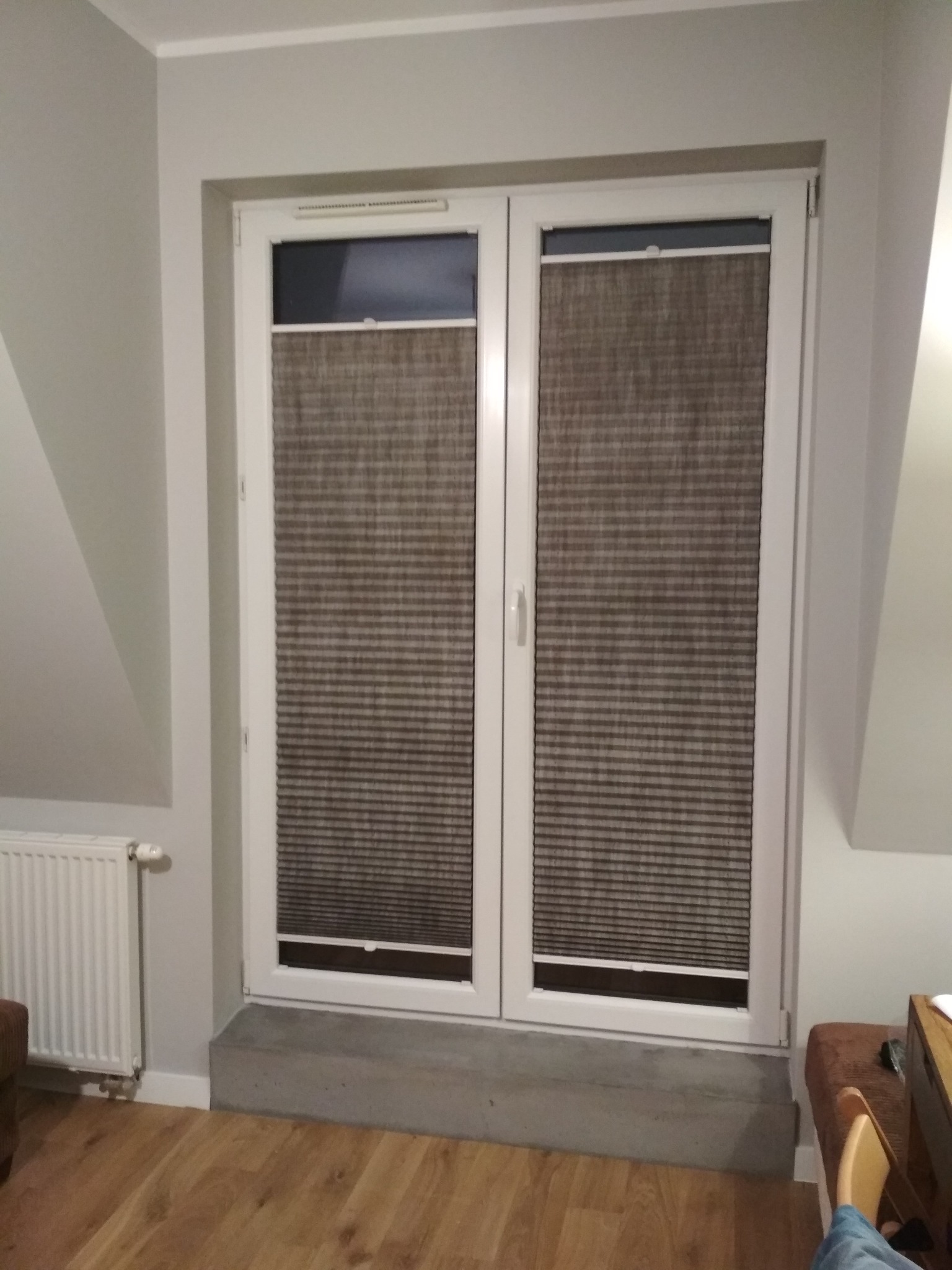 Dwu-skrzydłowe okno z białymi ramami, zabezpieczone plisowanymi żaluzjami w odcieniach szarości, zamontowane w pokoju na poddaszu z grzejnikiem.