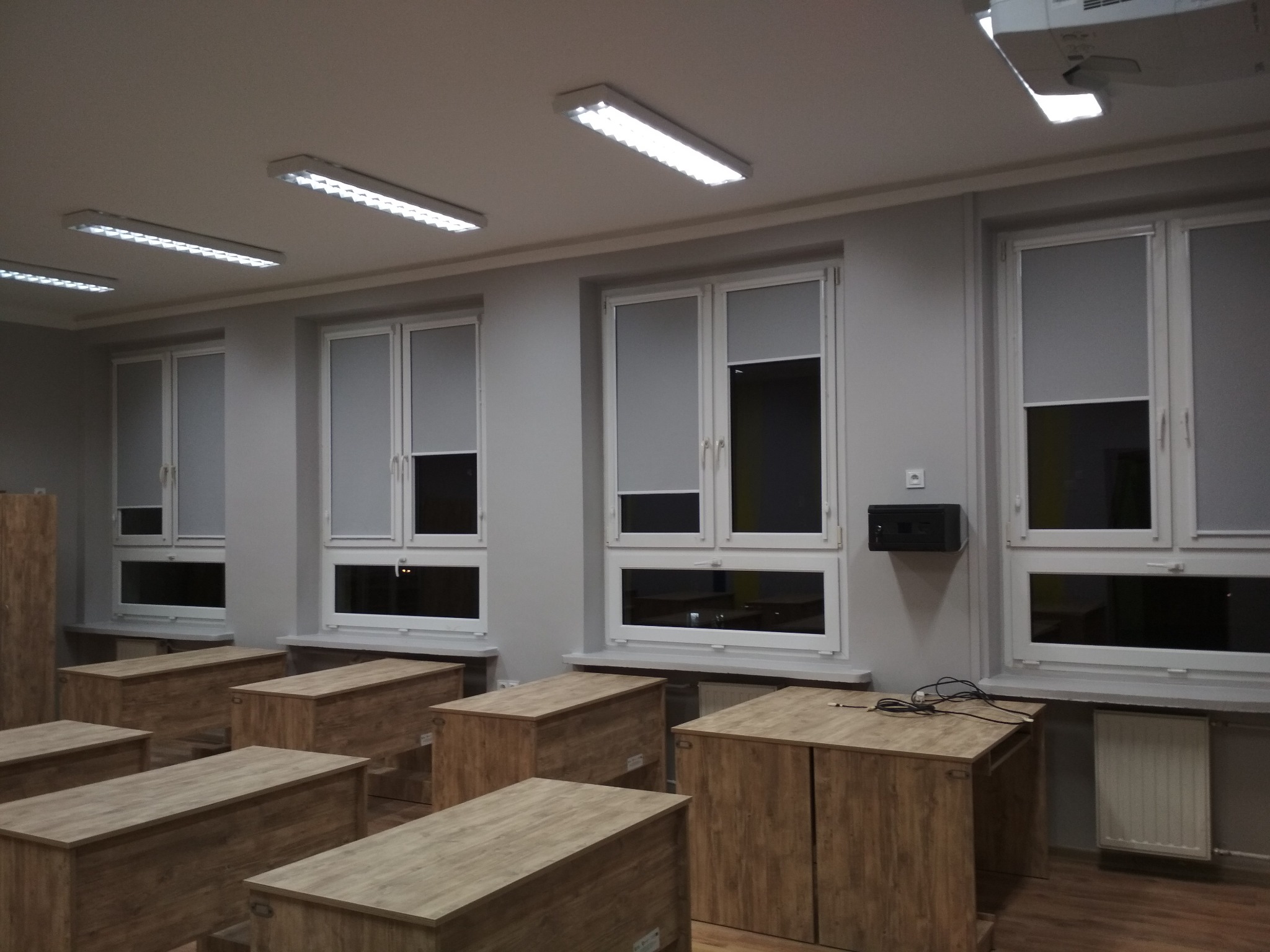Wnętrze klasy z roletami materiałowymi w kasecie, zamontowanymi w oknach PCV, widoczne drewniane szafki i projektor na suficie.