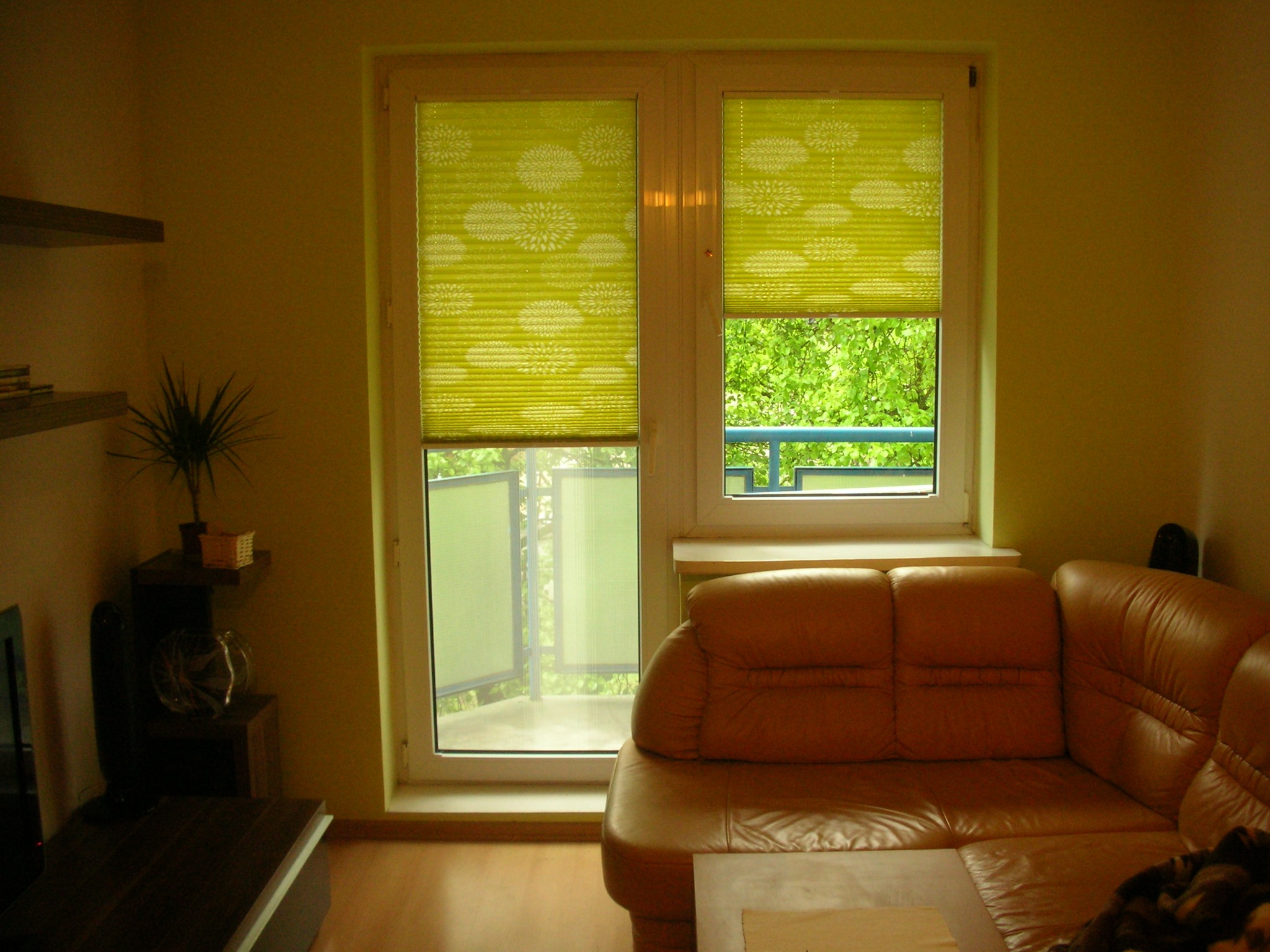 Wnętrze pokoju z oknem balkonowym, na którym zamontowane są zielone plisy z białym wzorem kwiatowym, częściowo zasłaniające widok na balkon i zieleń.