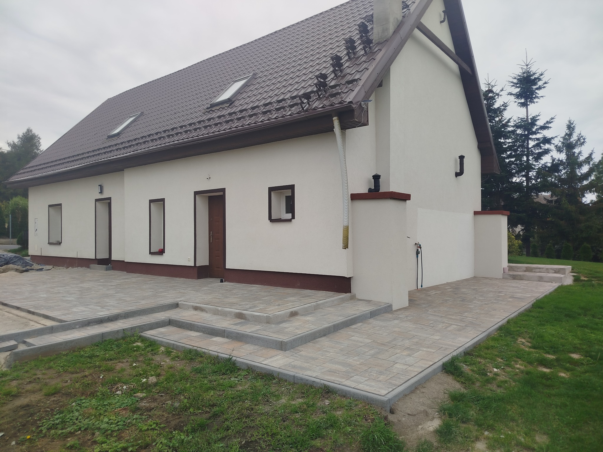 Jasna elewacja domu z brązowym dachem i nową, ułożoną kostką brukową na podjeździe i schodach wejściowych. Widok z poziomu gruntu, pochmurne niebo.