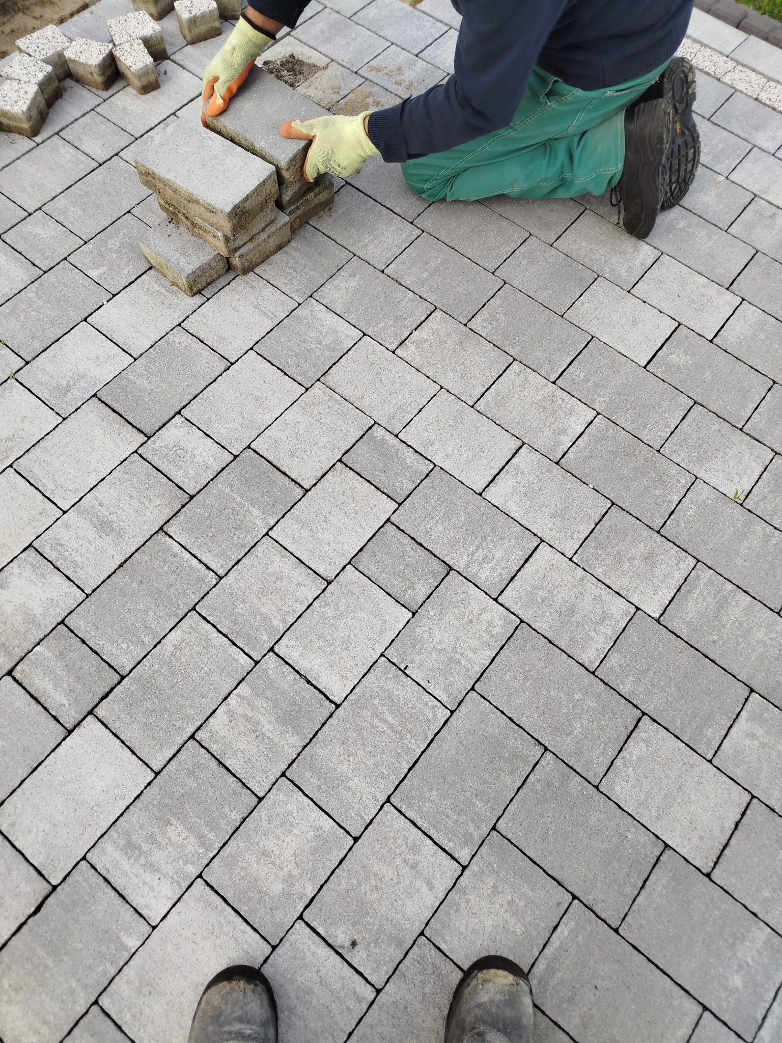Układanie szarej kostki brukowej w jodełkę. Widoczne dłonie w rękawicach, stos kostki i fragment buta. Ujęcie z góry, ukośne linie wzoru.