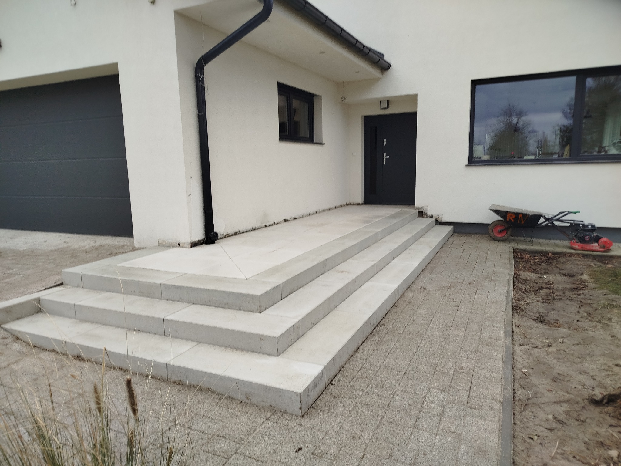 Nowoczesne schody wejściowe z jasnych płyt, prowadzące do domu z szarą kostką brukową. W tle widać taczkę i fragment ogrodu.