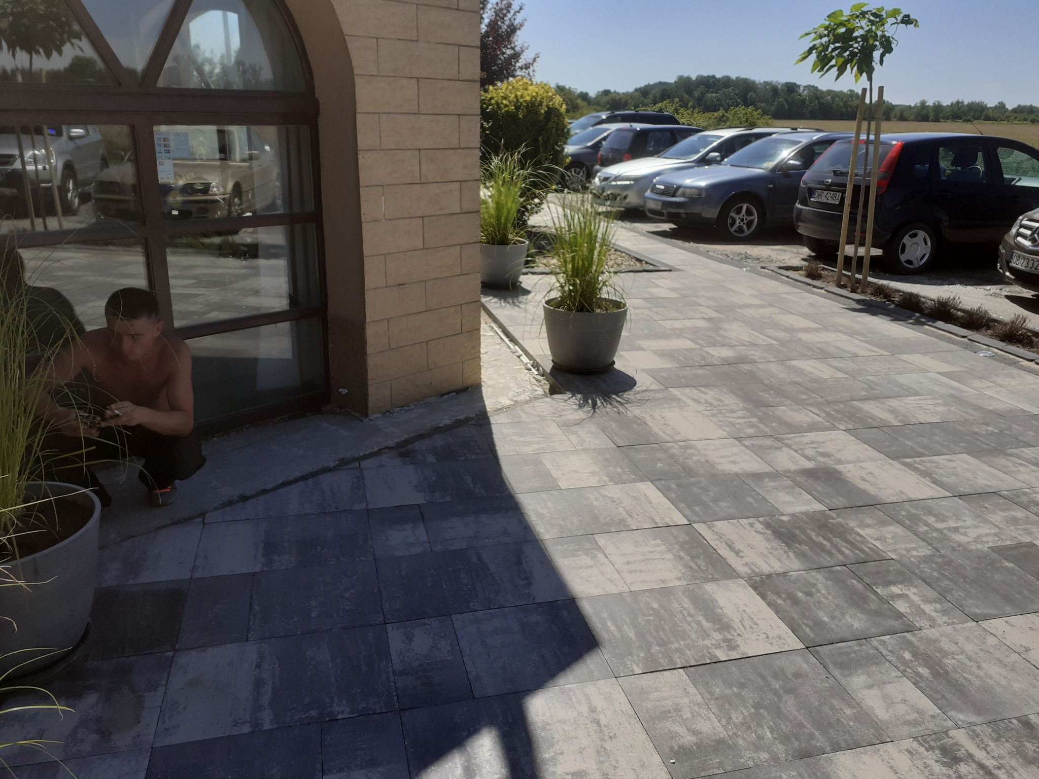Szara kostka brukowa ułożona na parkingu, z donicami i młodym drzewkiem. W tle zaparkowane samochody i budynek z dużym oknem łukowym, w którym odbija się otoczenie.