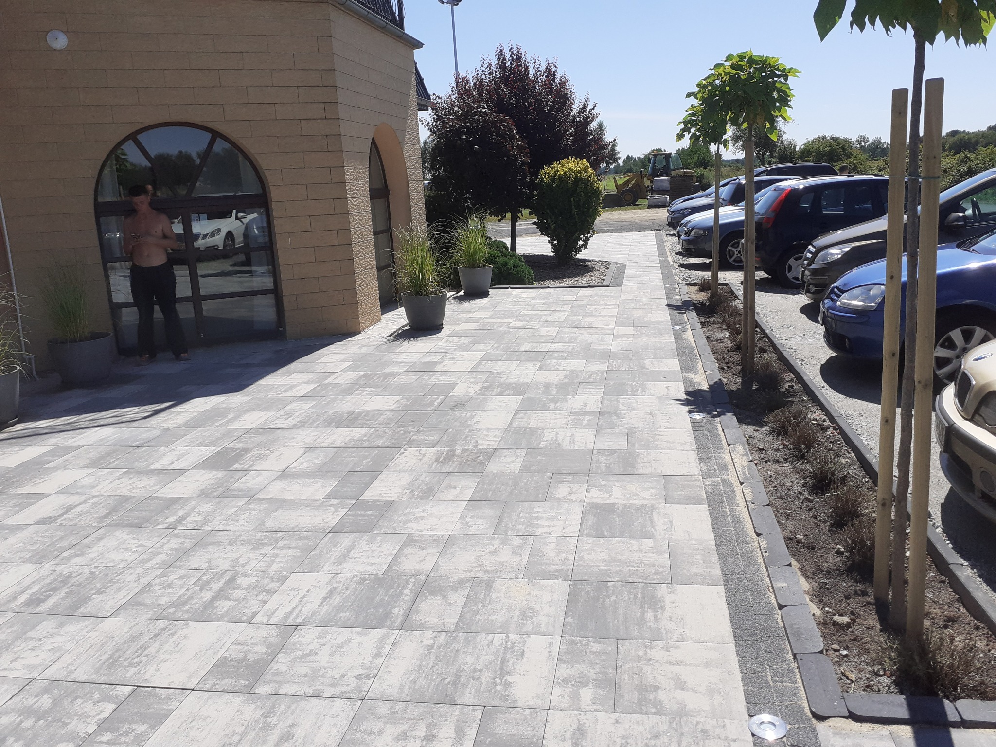 Ułożona kostka brukowa w odcieniach szarości prowadząca do budynku z oknem łukowym, obok zaparkowane samochody i drzewa. Widoczny fragment placu budowy w tle.
