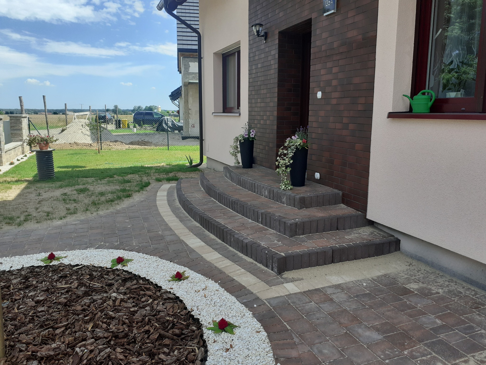 Kaskadowe schody z kostki brukowej prowadzą do domu. Dekoracyjny klomb z korą i białym żwirem, ozdobiony czerwonymi kwiatami. Estetyczne wykończenie posesji.
