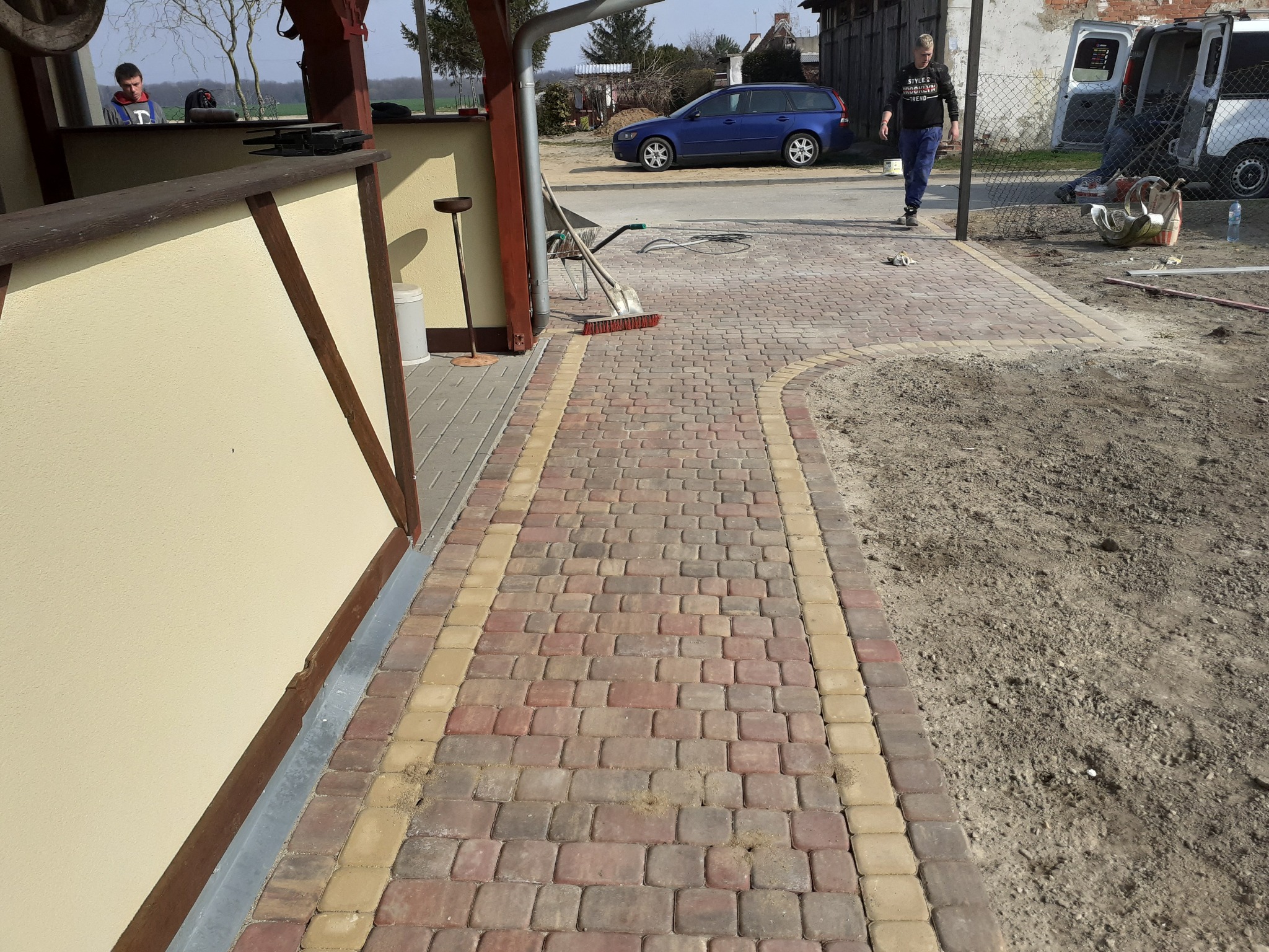 Ścieżka z kostki brukowej w odcieniach czerwieni i beżu, z widocznym fragmentem budynku i osobą w tle. Ujęcie z perspektywy chodnika, w słoneczny dzień.