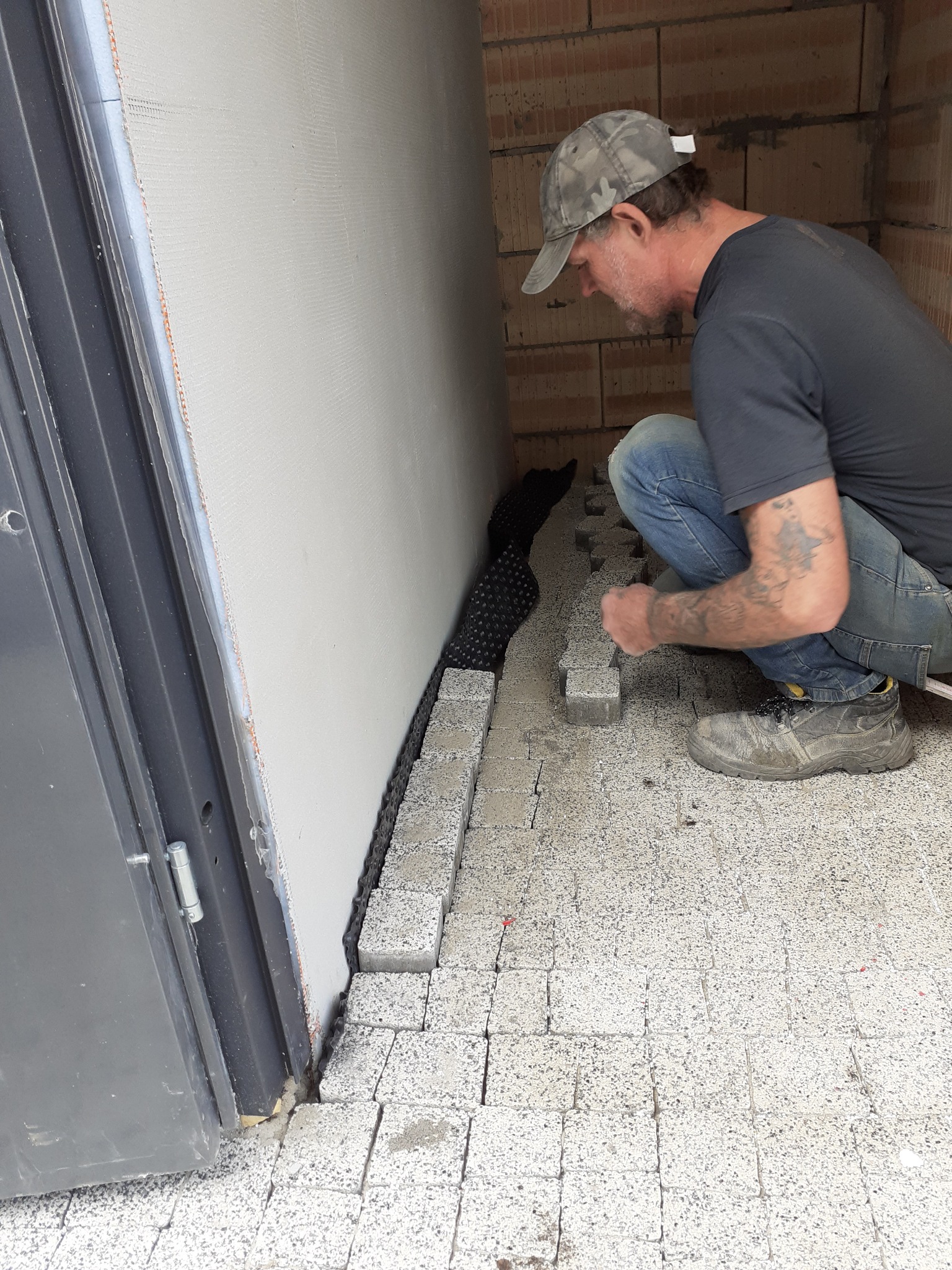 Mężczyzna w czapce układa szare kostki obok ściany z izolacją i czarnej folii drenażowej. Widoczny fragment ceglanej ściany i drzwi.
