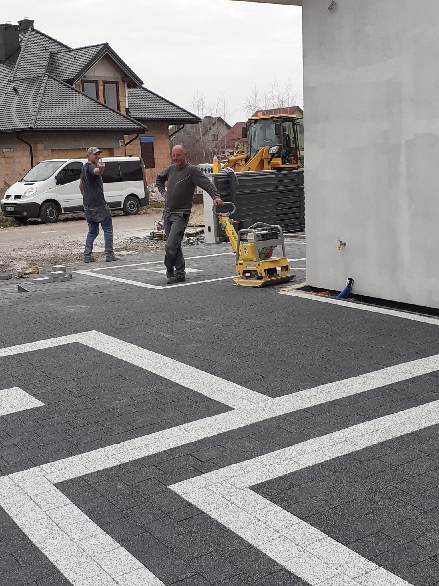 Ukończona nawierzchnia z kostki brukowej w geometryczny wzór, pracownik z zagęszczarką, w tle budynek i koparka. Widok z góry.