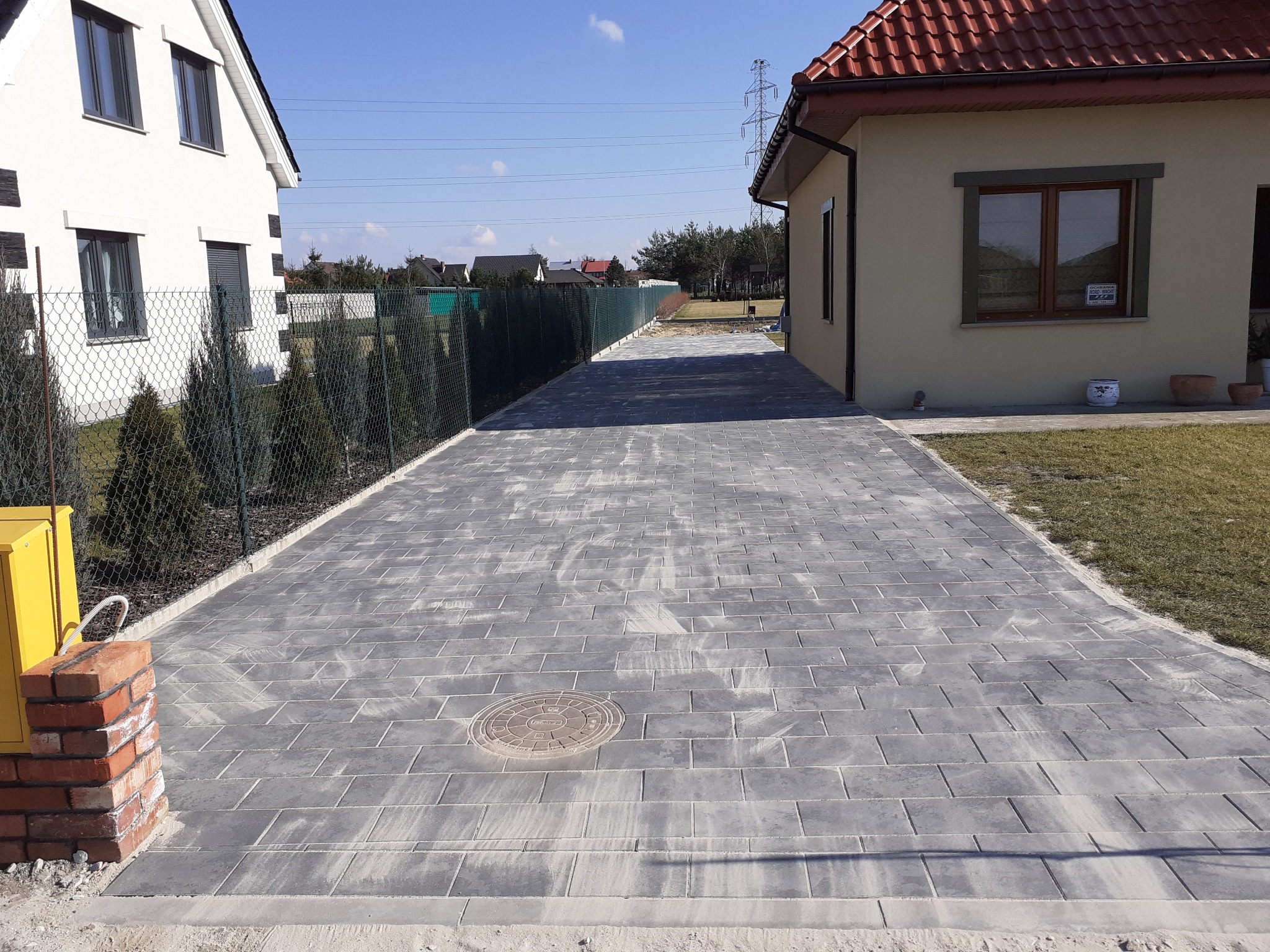 Długa, szara ścieżka z kostki brukowej prowadząca między domami, z widoczną studzienką kanalizacyjną i żółtą skrzynką gazową po lewej stronie.