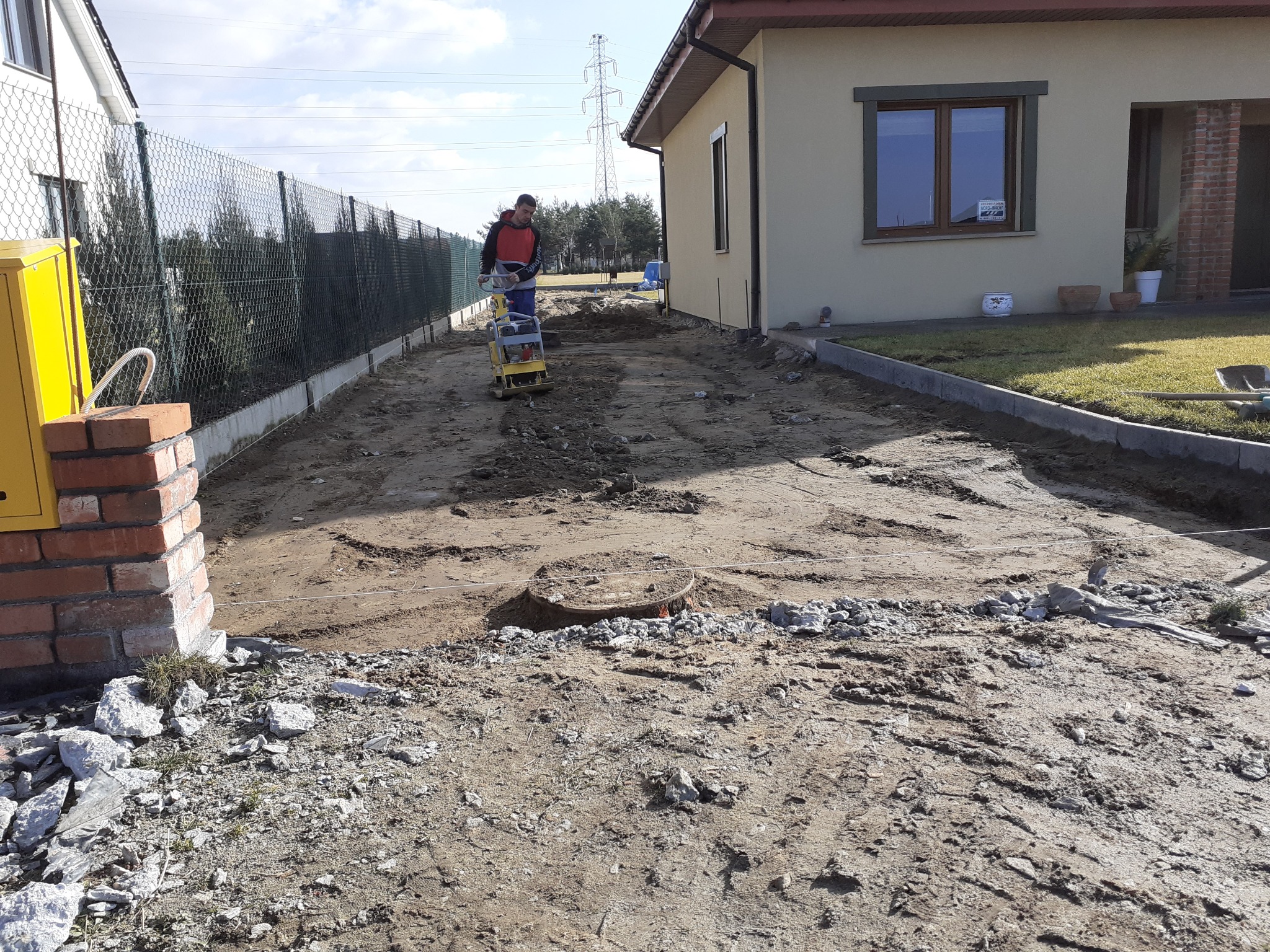 Prace przygotowawcze pod kostkę brukową: ubijanie ziemi zagęszczarką wzdłuż ogrodzenia i domu. Widoczny żółty gazomierz i krawężniki.