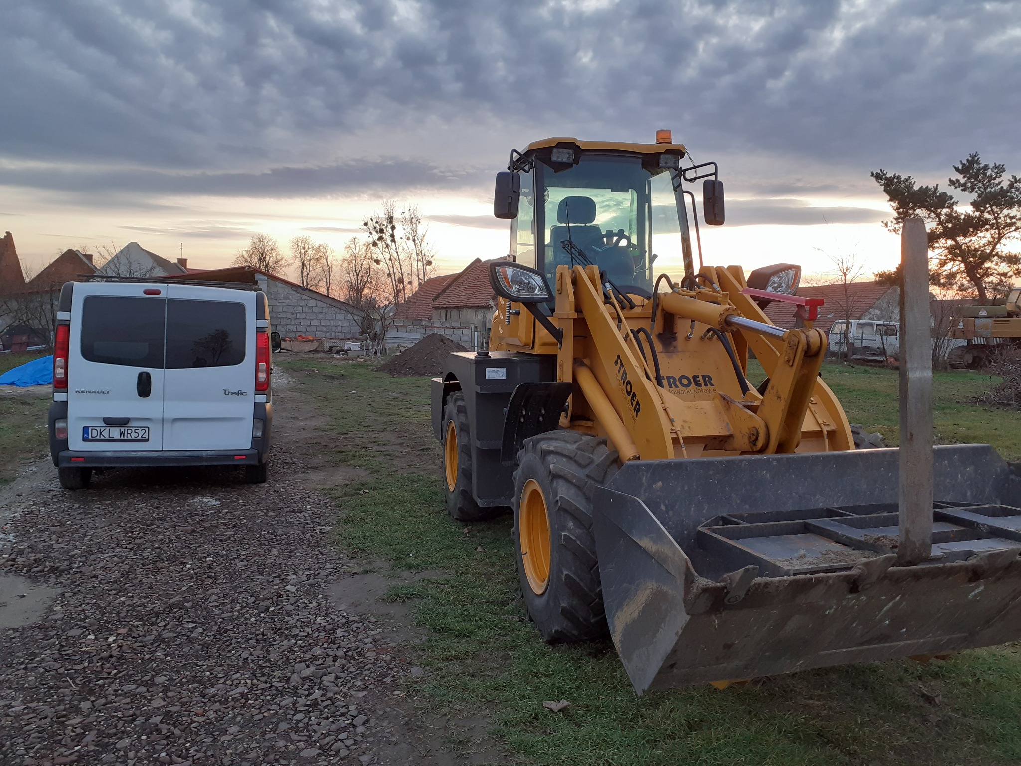 Żółta ładowarka STROER na trawiastym terenie, z białym busem Renault w tle i pochmurnym niebem o zmierzchu. Widoczne domy w oddali.
