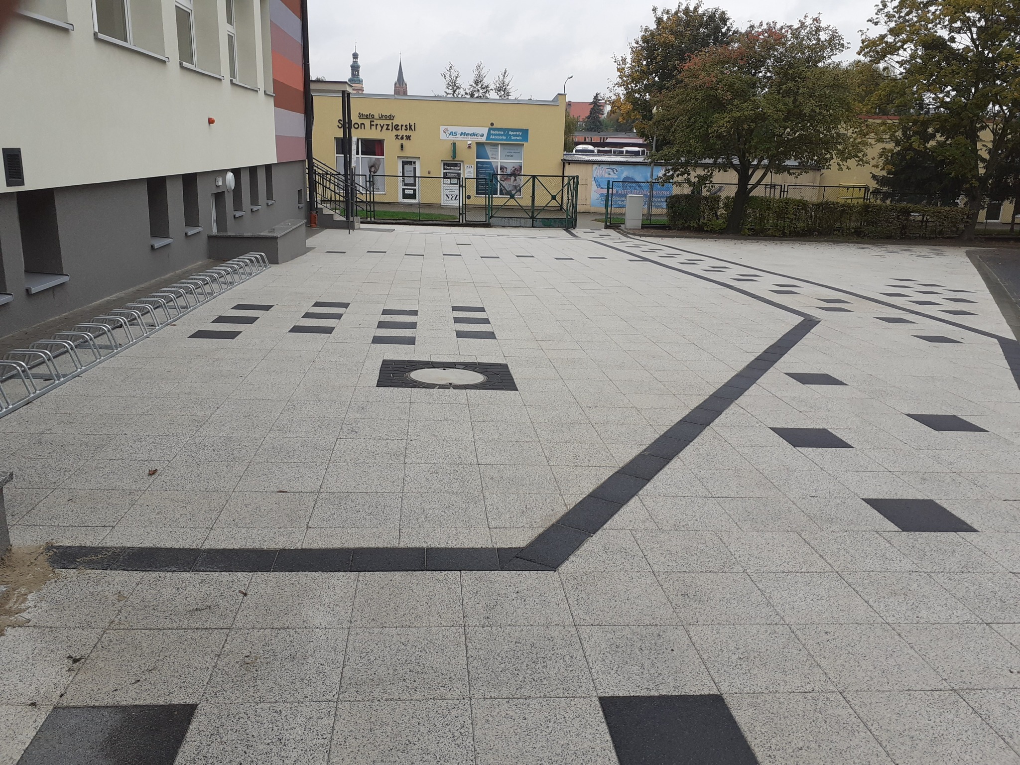 Szara kostka brukowa z ciemnymi, kwadratowymi wstawkami i liniami tworzącymi wzór na placu przed budynkiem z żółtą elewacją i stojakami na rowery.