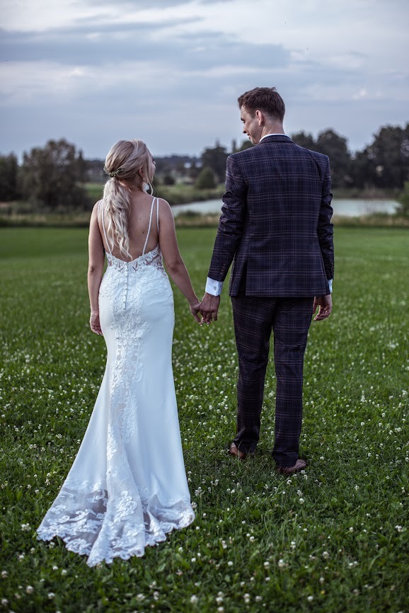 Para młoda, trzymając się za ręce, spaceruje po łące w kierunku stawu; panna młoda w białej sukni z koronkowymi zdobieniami na plecach i trenie, pan młody w ciemnym garniturze w kratę.