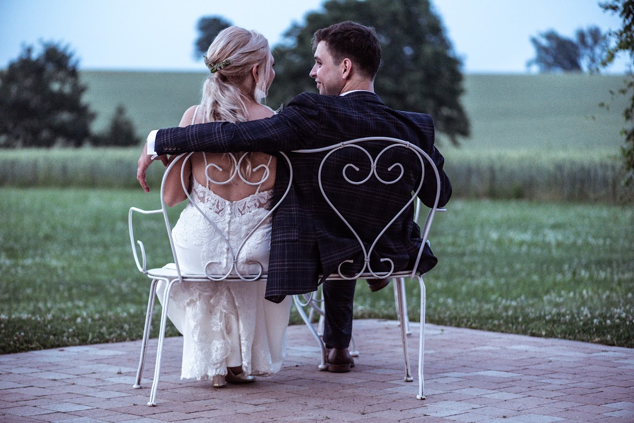 Para młoda siedzi tyłem na białych krzesłach z motywem serca, spoglądając na siebie na tle zielonego pola i drzew