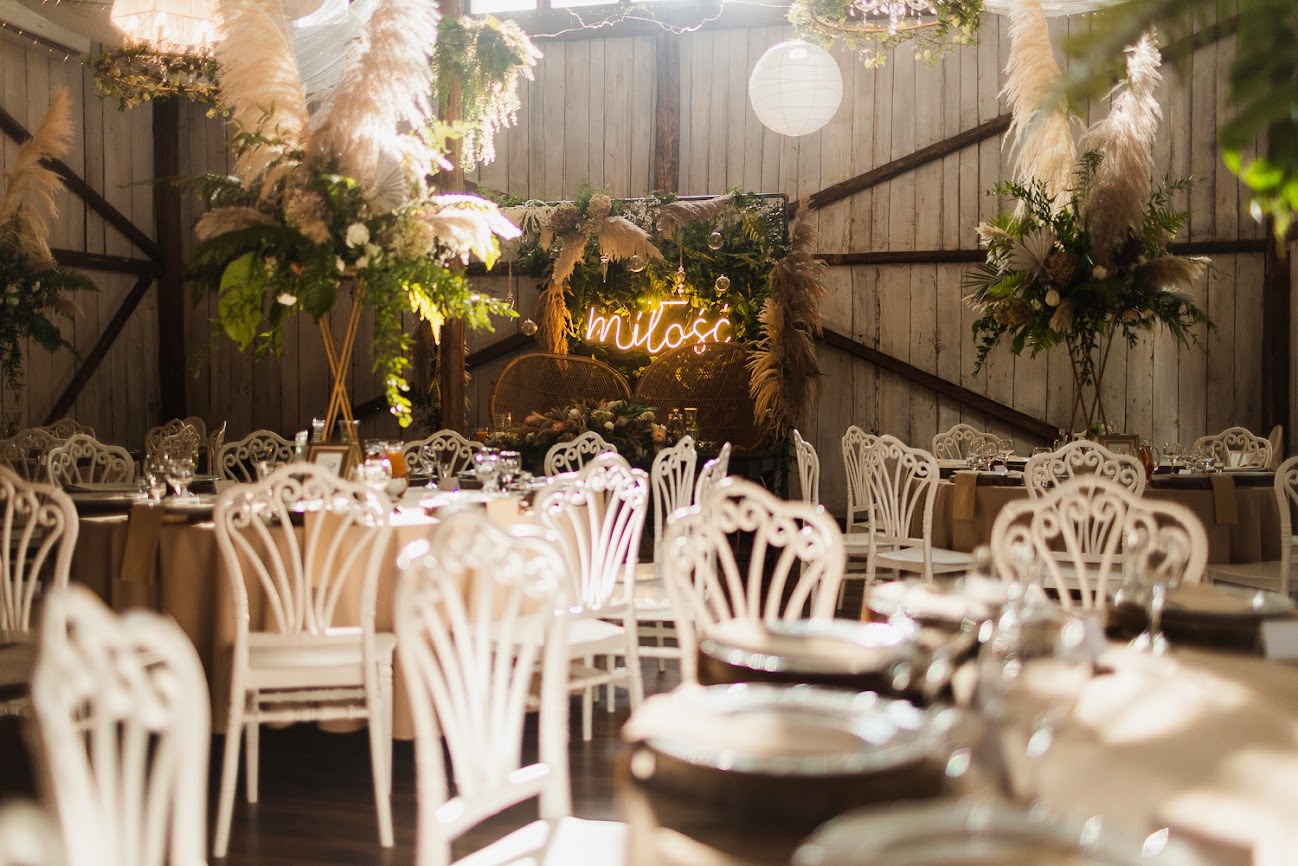 Sala weselna z dekoracjami: białe krzesła Chiavari, beżowe obrusy, kompozycje kwiatowe z trawą pampasową, neon 'Miłość' na tle drewnianej ściany.