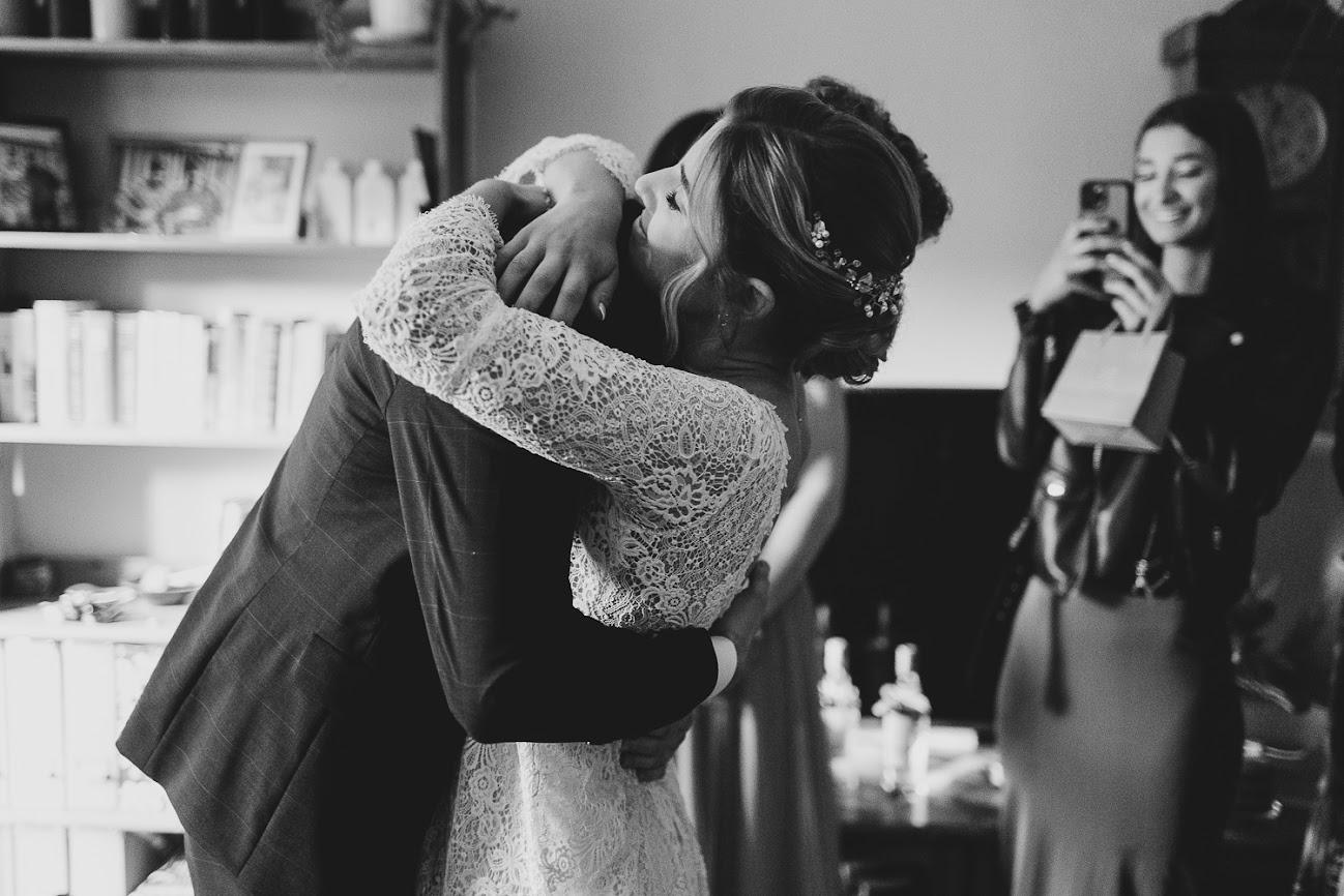 Czarno-białe zdjęcie obejmującej się pary młodej, panna młoda w koronkowej sukni i ozdobą we włosach, gość weselny robiący zdjęcie telefonem w tle.