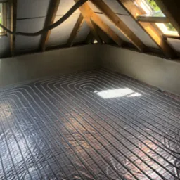 Rozłożone rury ogrzewania podłogowego na folii izolacyjnej z czerwoną siatką, widoczne poddasze z oknem dachowym i drewnianą konstrukcją.