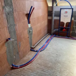 Surowe wnętrze łazienki w trakcie instalacji: widoczne czerwone i niebieskie rury instalacji wodnej prowadzone po podłodze, częściowo zamurowane w ścianie z cegły, stelaż podtynkowy WC marki Grohe...