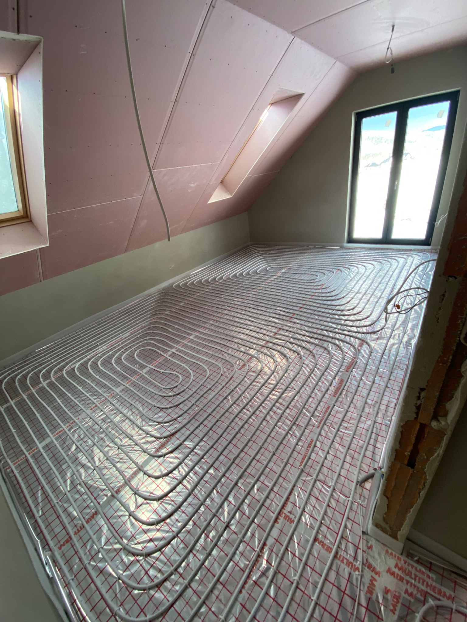 Białe rury ogrzewania podłogowego ułożone spiralnie na folii izolacyjnej w pokoju na poddaszu ze skośnym sufitem i oknami dachowymi, instalacja w trakcie realizacji.