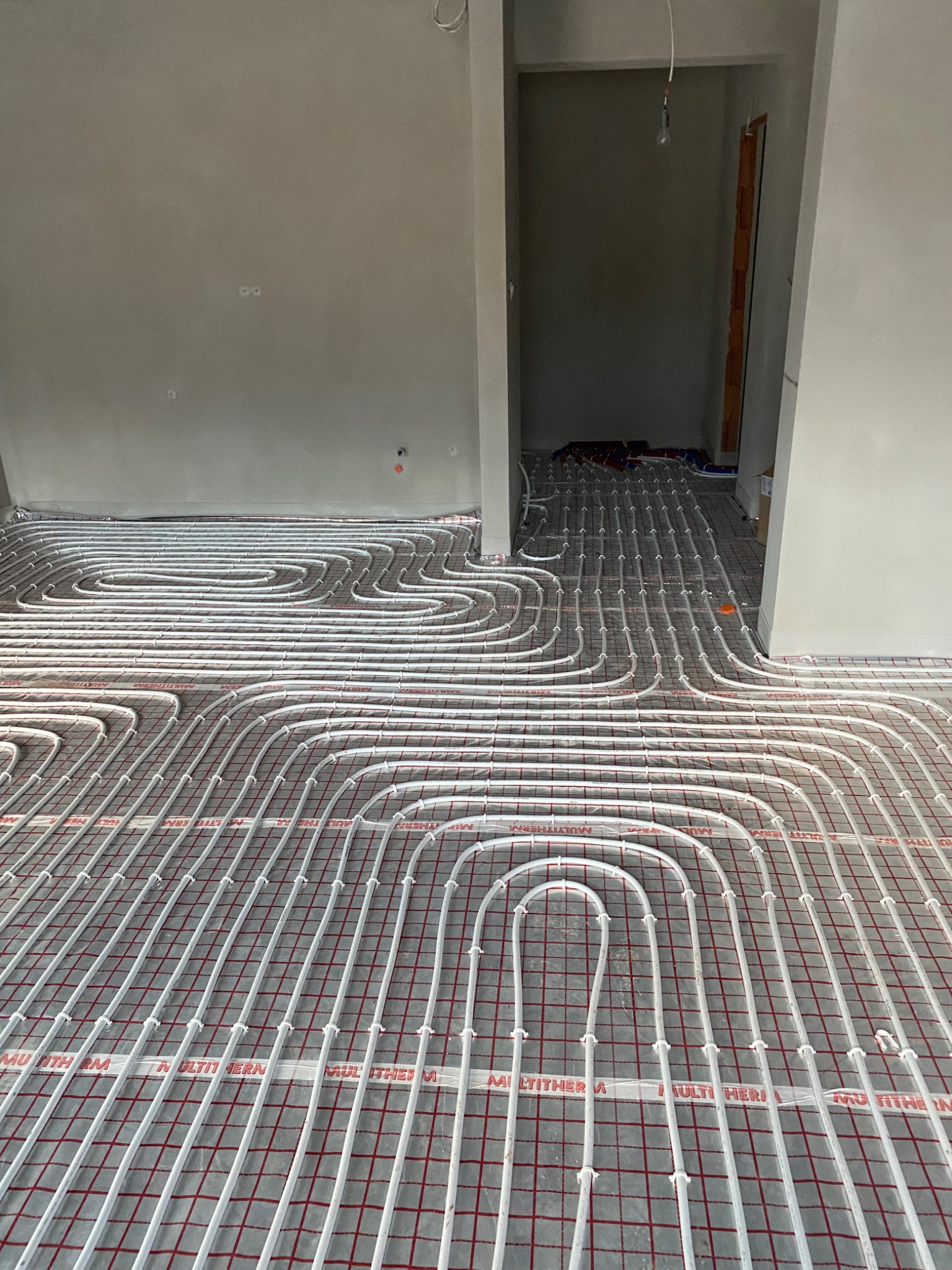 Rozłożona instalacja ogrzewania podłogowego na siatce montażowej, widoczne białe rury MULTITHERM przed wylewką, zarys pomieszczenia w tle.